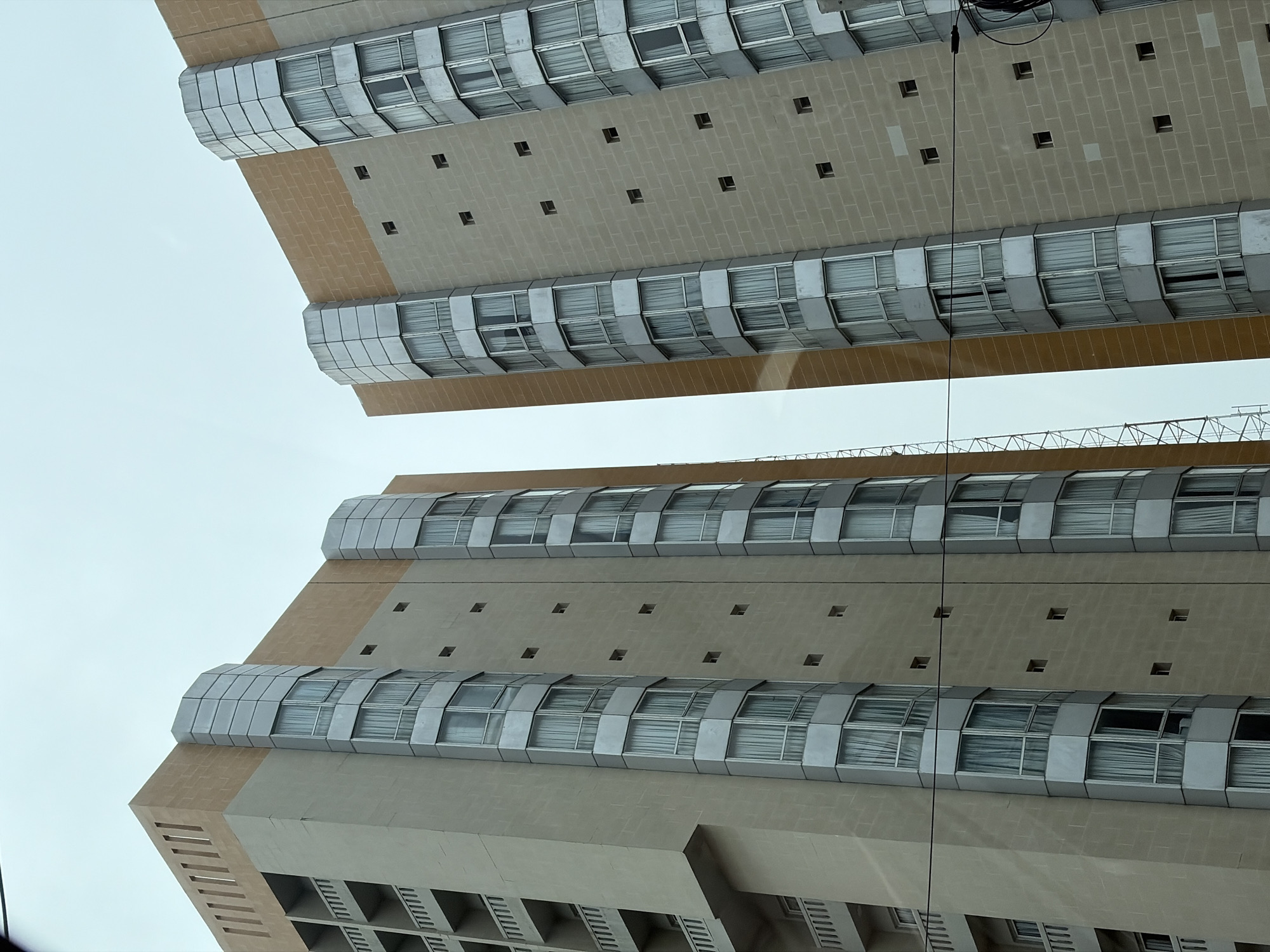 The image shows the exterior of a modern building in Falomo, Nigeria. The building has a beige facade with multiple tiers of windows that curve outwards. There are small square openings on the facade between the window tiers. The sky is visible in the background, suggesting an upward-looking perspective. A dark cable is visible near the top of the frame.