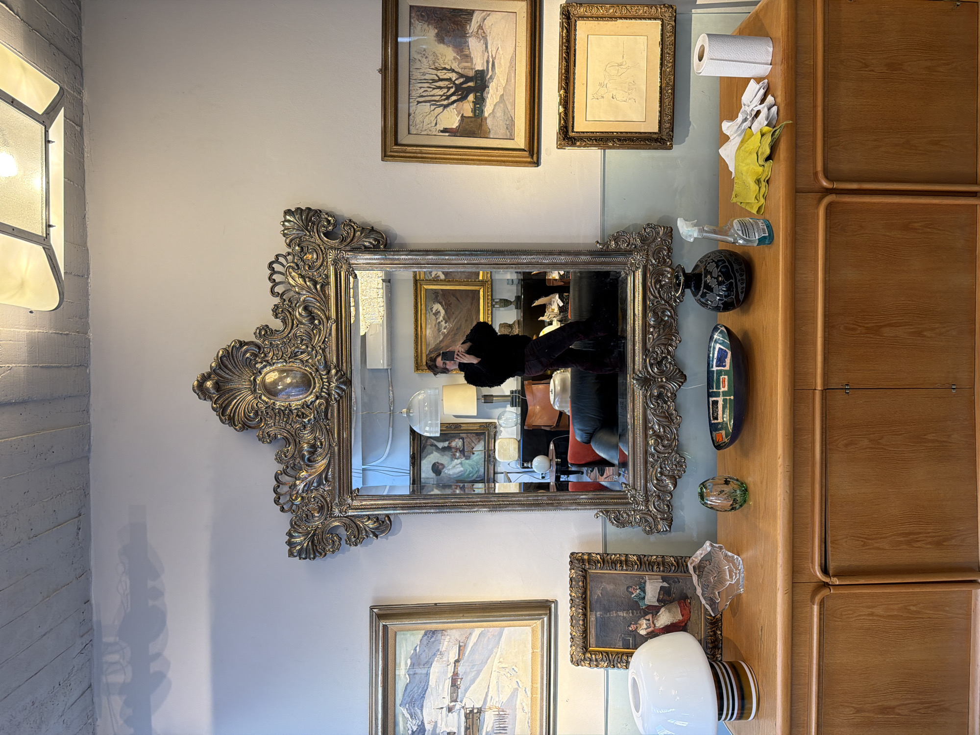 The image depicts an interior wall in Buenos Aires, Argentina, adorned with artwork and decorative items. Dominating the scene is a large, ornate, antique-style mirror. Above the mirror are two framed pictures, while below is another framed picture. To the right of the mirror is a wooden cabinet, on which are placed cleaning supplies (paper towels, spray bottle, and yellow cloth), and several decorative bowls/vases. A white lamp sits at the bottom corner of the wall. The reflection in the mirror captures a person holding a phone, presumably taking the photo, along with elements of the room behind them, including furniture and additional artwork.