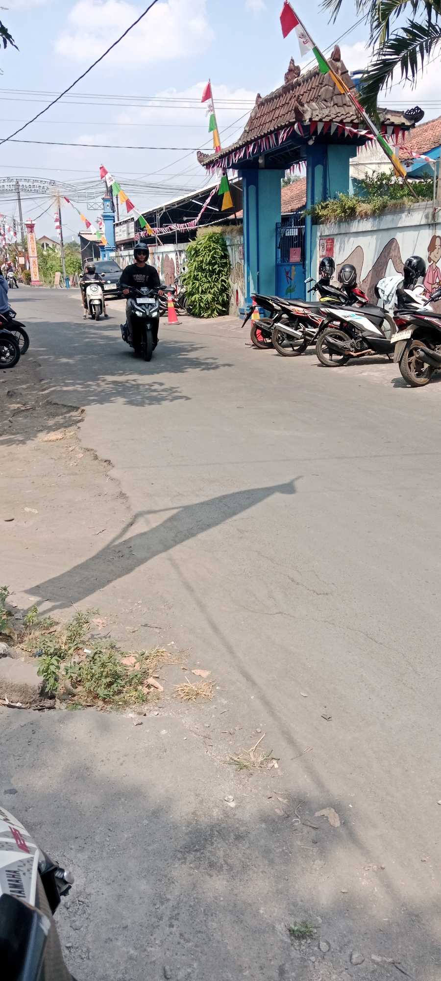 A person rides a black scooter on a paved road in Purwomartani, Indonesia, heading towards an entrance gate decorated with Indonesian flags. The gate, adorned with traditional roof tiles, is painted a bright blue. A mural featuring cartoon figures is visible on the wall to the right of the gate. Several other scooters are parked to the right of the gate. The sky is blue with scattered clouds. Shadows from a person and a scooter are cast on the road.