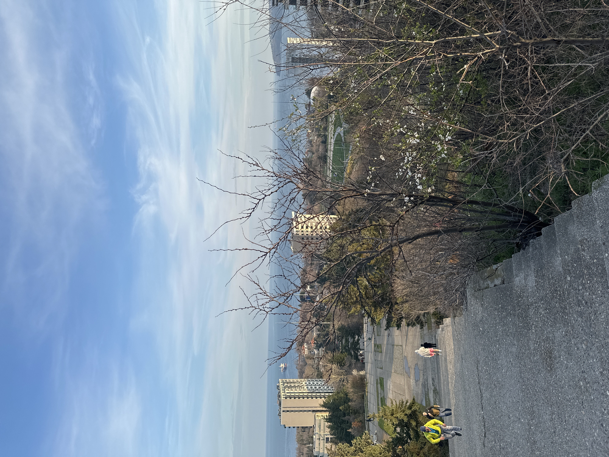 The image shows a view from a high vantage point in Varna, Bulgaria. The sky is blue with scattered clouds. In the distance is the Black Sea. In the middle ground, there's a mix of vegetation and city buildings. On the right, the foreground shows a stone staircase with people walking down it. One person is wearing a bright yellow jacket. A building can be seen in the center of the photo that is on the waterfront.