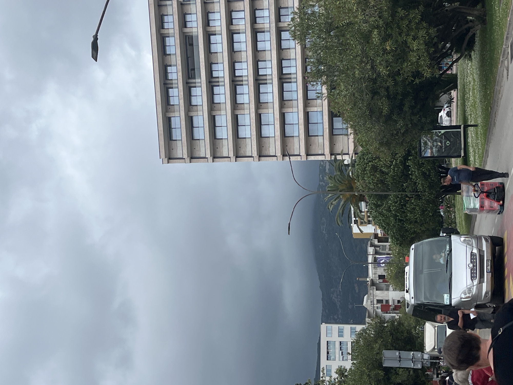 The image captures a street scene in Budva, Montenegro, under an overcast sky. Tall buildings dominate the background, with a modern building featuring rows of windows on the left, and residential buildings visible in the lower right. In the foreground, a white van is partially visible, surrounded by people, some of whom appear to be tourists. A person pulls a red cart across the street. Lush green trees and vegetation are interspersed throughout the scene. A billboard stands on the side of the road. Mountains loom in the distance behind the town.