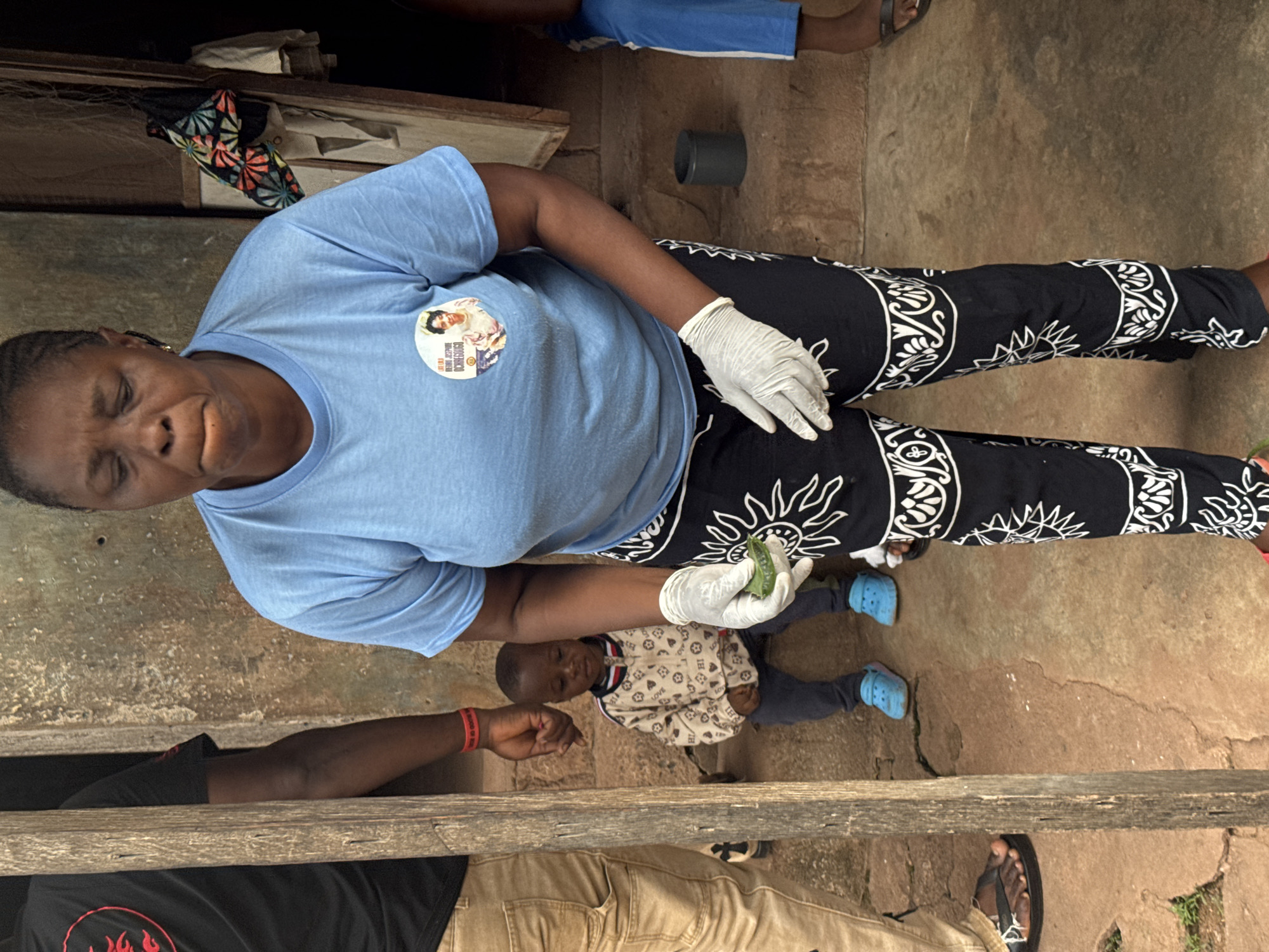 A Nigerian woman wearing a blue t-shirt and black pants with white sun-like patterns is standing and holding a green piece of okra. She is wearing white disposable gloves. A young child, wearing a patterned jacket and blue shoes, is sitting on the ground to her left. Another person, wearing a black t-shirt and tan pants, is standing to the far left, with a red wristband. The scene takes place in an outdoor setting with a dirt floor and a rough, textured wall in the background. This appears to be an urban setting, potentially in Enugu, Nigeria.