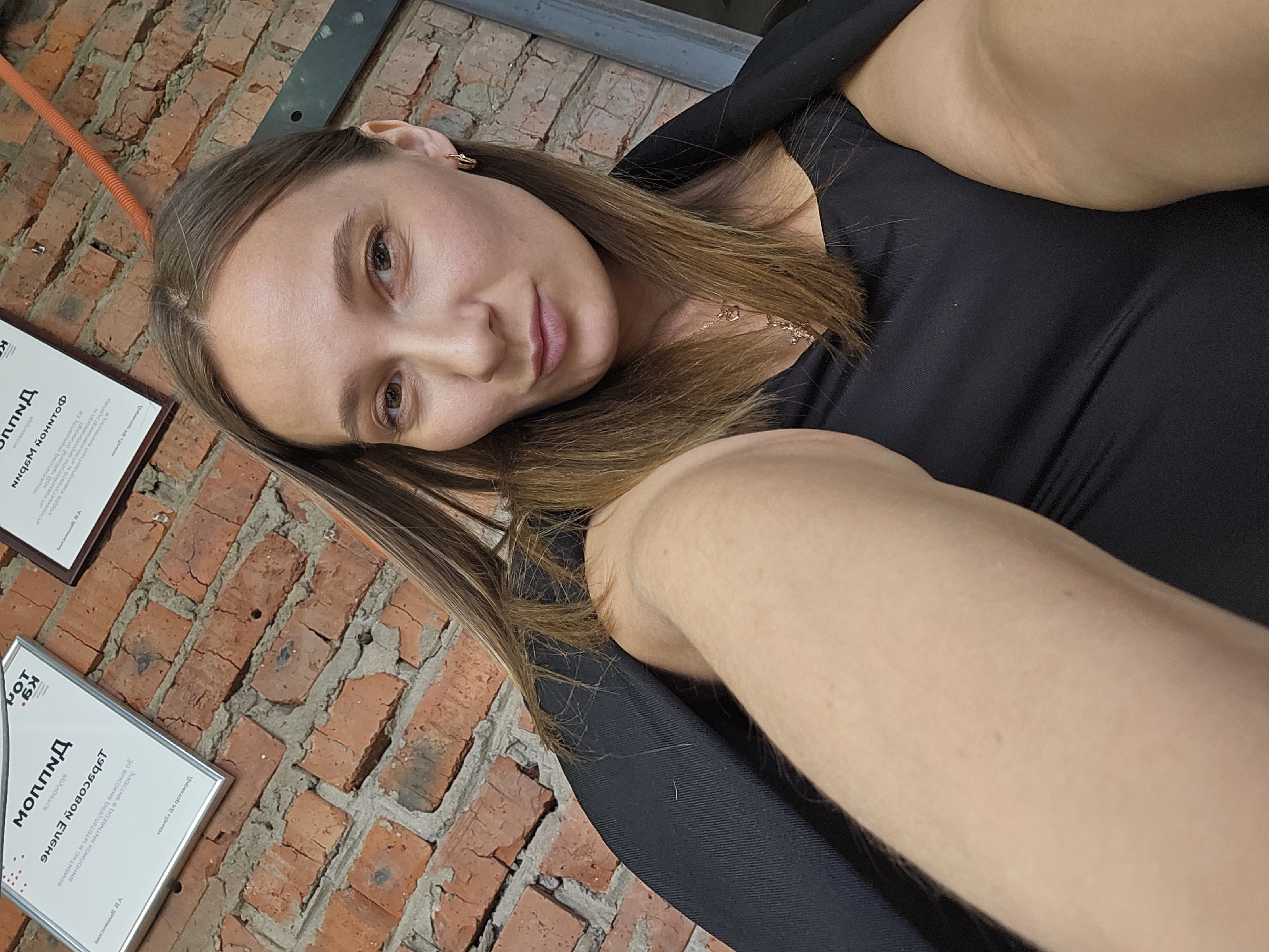 A woman is featured in a close-up selfie, looking directly at the camera with a calm expression and a faint smile. She has medium-length brown hair, parted in the middle, and is wearing a black top or dress, adorned with a delicate gold necklace and a small gold earring in her visible ear. Her extended arm, holding the camera, is partially visible in the foreground.
The background consists of a rustic, exposed reddish-brown brick wall with grey mortar. An orange cable or pipe runs diagonally across the upper left of the wall, and a dark metal beam is visible in the upper right. Two framed certificates or documents hang on the brick wall.
The visible Cyrillic text on these documents includes "МОППИД" (Moppid), potentially a brand or organization name. One certificate displays a "К9" logo with a paw print, and mentions "ЕВГЕНИЙ ЗАБОБОН" (Evgeniy Zabobon), "УЧАСТНИК" (Participant), "ВЫСТАВКИ" (Exhibition), and "КИНОЛОГ" (Canine specialist/Dog handler). This suggests the documents are certificates of participation or achievement in dog-related events or training.
The setting appears to be an indoor venue, possibly a cafe, business, or training facility in Chaikovsky, Russia, given the Cyrillic text on the documents. The overall impression is one of a casual, personal moment captured against an interesting textural background.