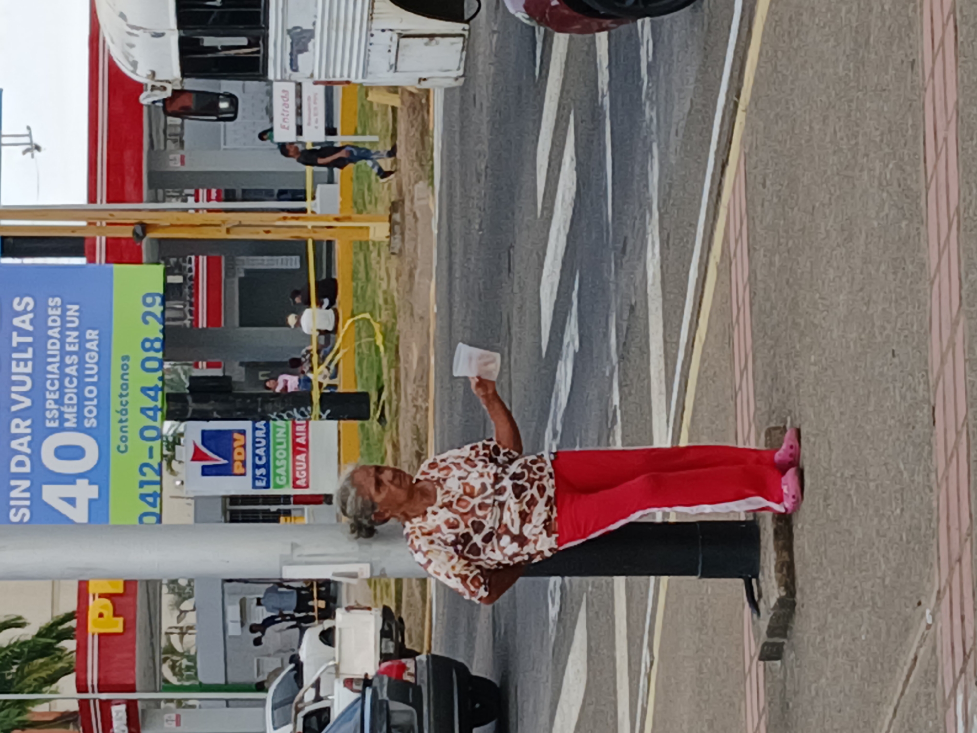 The image shows an elderly woman standing on the sidewalk in Guayana City, Venezuela. She has grey hair pulled back in a bun, wears a patterned top and red pants, and pink sandals. She is holding a white object in her raised right hand.
She stands next to a utility pole on the sidewalk that runs along a busy road. Cars are visible in the foreground. In the background, there is a gas station labeled "PDV E/S CAURA," along with signs advertising gas, water, and air. A large blue sign displays the text "SIN DAR VUELTAS 40" followed by "ESPECIALIDADES MEDICAS EN UN SOLO LUGAR" and a phone number. People are moving around the gas station area.