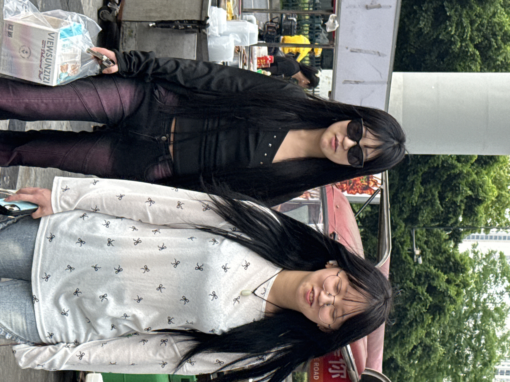 Two young women stand next to each other in what appears to be a public space in the Chongqing High-tech Zone, China. The woman in the foreground wears a white long-sleeved shirt with a small bow pattern, blue jeans, and circular eyeglasses. She has long, dark hair. The other woman, slightly behind, is dressed in a black top, black pants with a purple hue, and sunglasses. She also has long, dark hair. A box with text ("VIEWSUNZIZI") is visible to the left of the women. A street sign with Chinese characters is partially visible on the bottom right. Trees and a building can be seen in the background.