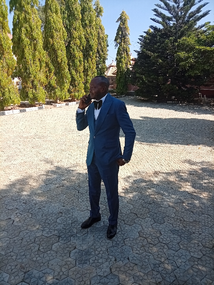 An adult male stands outdoors on a grey hexagonal interlocking paved surface under a clear blue sky. He wears a dark blue three-piece suit, a white shirt, a black bow tie, black dress shoes, and a silver-toned wrist watch on his left arm. He holds a dark-colored smartphone to his right ear with his right hand, appearing to be in a conversation with his mouth slightly ajar. His left hand is in his left trouser pocket. In the mid-ground, a row of tall, slender evergreen trees with dense green foliage stands behind him, bordered by a low white curb. To the right of the frame, a large, broad-canopied evergreen tree is visible. Portions of light-colored buildings with red-tiled roofs and decorative architectural features are partially obscured by the trees in the background. Bright sunlight casts distinct shadows across the paved area. The scene is situated in the city of Lankan, Nigeria.
