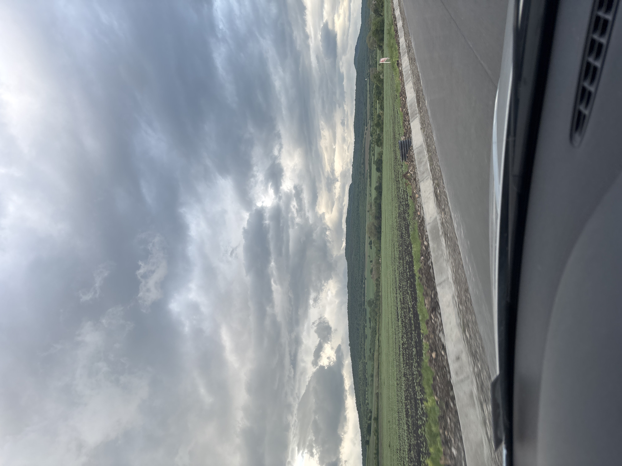 The image shows a view from inside a vehicle, possibly a car. The foreground is dominated by the vehicle's interior. Outside the vehicle, the landscape features a green field that extends towards a line of trees or low hills. A road runs parallel to the field. The sky is overcast with heavy cloud cover. There is no indication that this picture was taken in Belopoptsi, Bulgaria.