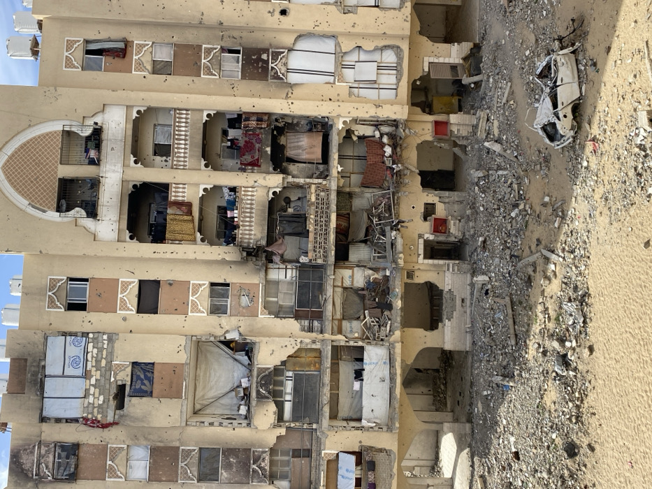 This image displays a severely damaged multi-story residential building located in Khan Yunis, Palestinian Territory, under clear daylight conditions. The light-colored building facade is extensively pockmarked with numerous small impact craters consistent with bullet holes or shrapnel, distributed across all visible floors. Large sections of the building's exterior walls are collapsed or torn away, exposing interior rooms and structural components. Balconies on multiple levels are partially destroyed or heavily compromised, with some still showing draped clothing. Many window frames are either absent, shattered, or sealed with makeshift materials such as metal sheeting and fabric. White water tanks are visible on the building's rooftop. The ground in front of the structure is covered in a dense layer of pulverized concrete, rubble, and sand. In the lower right foreground, a heavily mangled and crushed white four-door sedan, stripped of much of its bodywork, rests immobile amidst the debris. No individuals are visible in the scene.