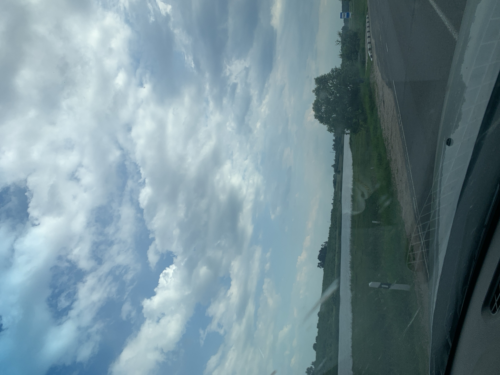 The image depicts a roadside view on a cloudy day. A paved road curves to the right, with a grassy embankment and a body of water on the left. Several trees are visible on the far bank of the water and further inland. A road sign is present on the right side of the road. The foreground shows the dashboard and windshield of a vehicle, suggesting the photo was taken from a car. The sky is filled with dramatic clouds. The location is identified as Слабада-Кучынскі сельскі Савет, Belarus.