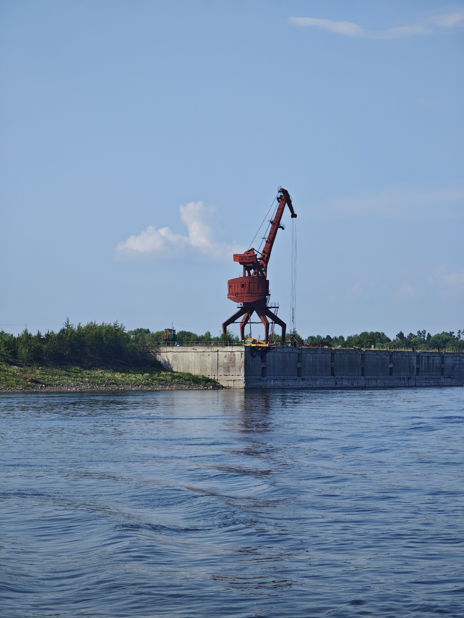 A red gantry crane stands on a concrete pier on the bank of a wide, blue river under a bright blue sky with a few scattered white clouds. The pier is lined with a concrete wall and a railing. Behind the pier, there is a line of green trees and bushes. The water in the foreground is rippled, suggesting a slight current or breeze. The scene appears to be an industrial waterfront, possibly a port or loading facility. There are no people visible in the image. The location is the city of Verkhnepashino, Russia.