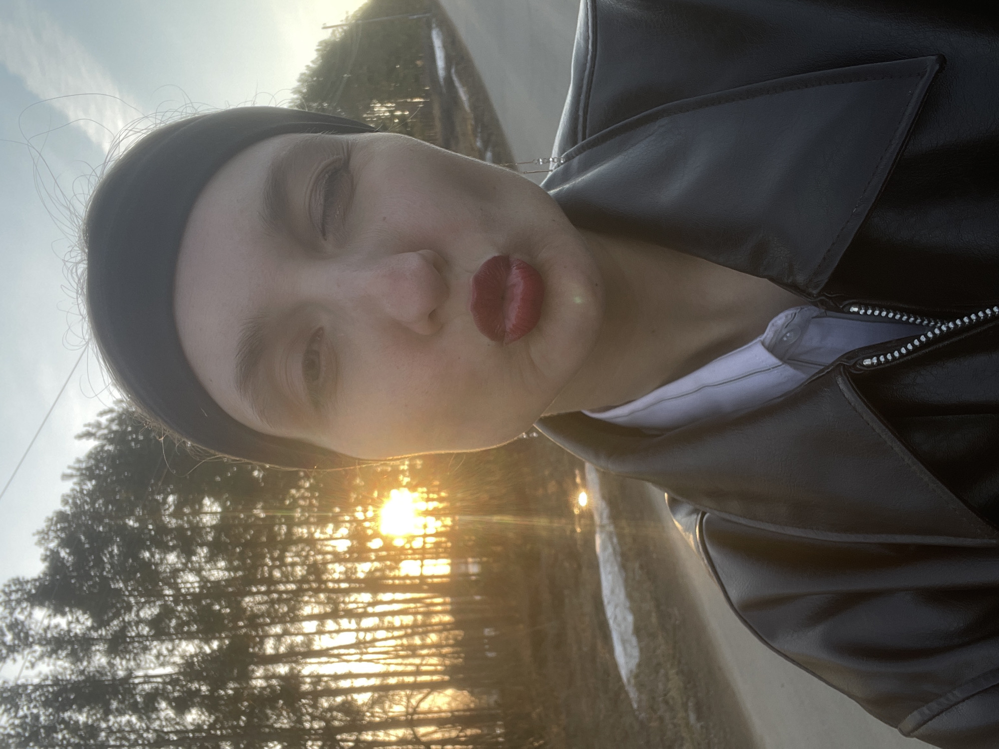 A close-up, tilted selfie of a person with fair skin and red lipstick is captured against a blurred background. The person has a dark headband and wears a black leather jacket over a white shirt. The background shows trees and a bright sunlight filtering through the foliage, with what appears to be a road or path visible. The location appears to be Podshivalovo, Russia.