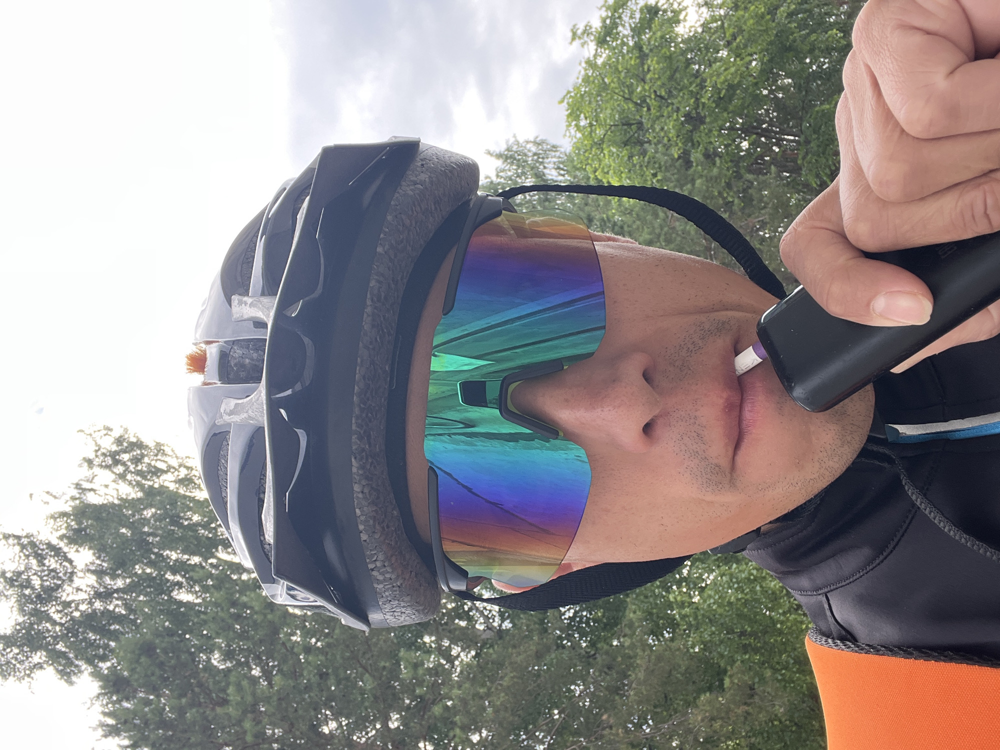 This media file captures a close-up, low-angle shot of a person wearing a black cycling helmet and iridescent, wrap-around sunglasses. The individual is holding a black electronic device, possibly a vape or e-cigarette, to their lips. The focus is on the person's face, particularly their mouth and the sunglasses which reflect a vibrant spectrum of colors. The background consists of out-of-focus green trees and a bright, overcast sky, suggesting an outdoor setting. The person appears to be engaged in an activity, likely cycling, given the helmet. The overall impression is a candid moment captured during an outdoor excursion, with the weather appearing fair but cloudy. There is no visible text in the image.