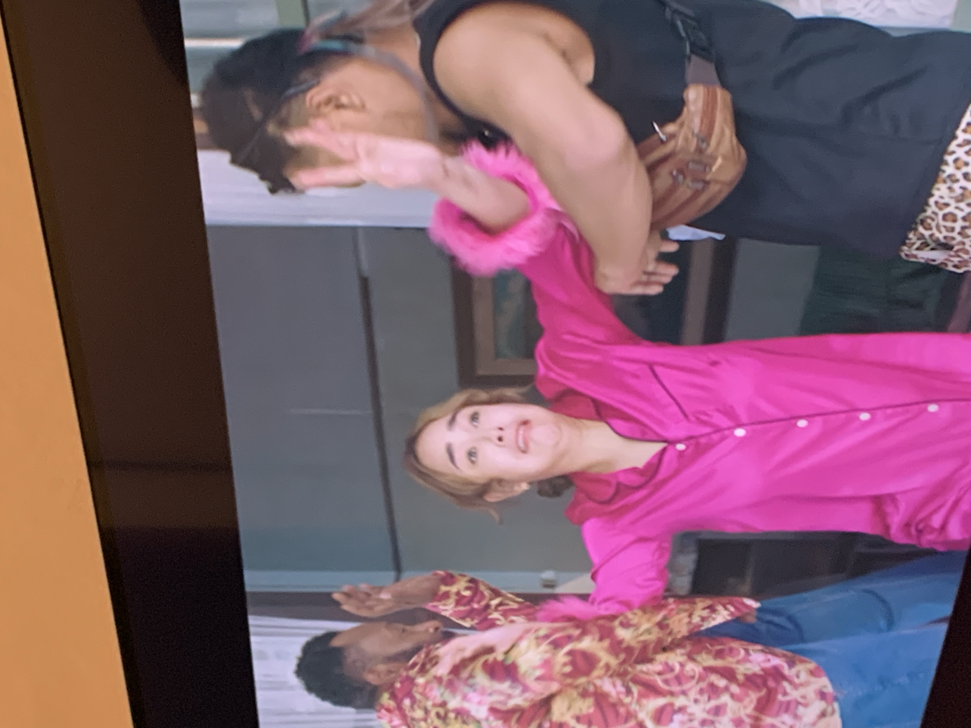 In the city of Kualadua, Indonesia, a scene unfolds on a television screen, depicting three individuals in what appears to be an indoor setting. A woman, wearing a bright pink pajama set, is positioned centrally, her face contorted in an expression of fear or distress. Her hands are raised, palms outward, in a defensive gesture. To her left, a man in a patterned red and gold shirt and blue pants also has his hands raised in a similar defensive posture, facing away from the camera. To the woman's right, another individual, partially obscured and facing away, is seen with one hand raised, fingers spread in a peace sign gesture. This individual is wearing a black tank top, a brown fanny pack, and leopard-print shorts. The background suggests an interior space with muted, possibly gray walls and what looks like a window. The image captures a moment of apparent conflict or tension between the subjects.