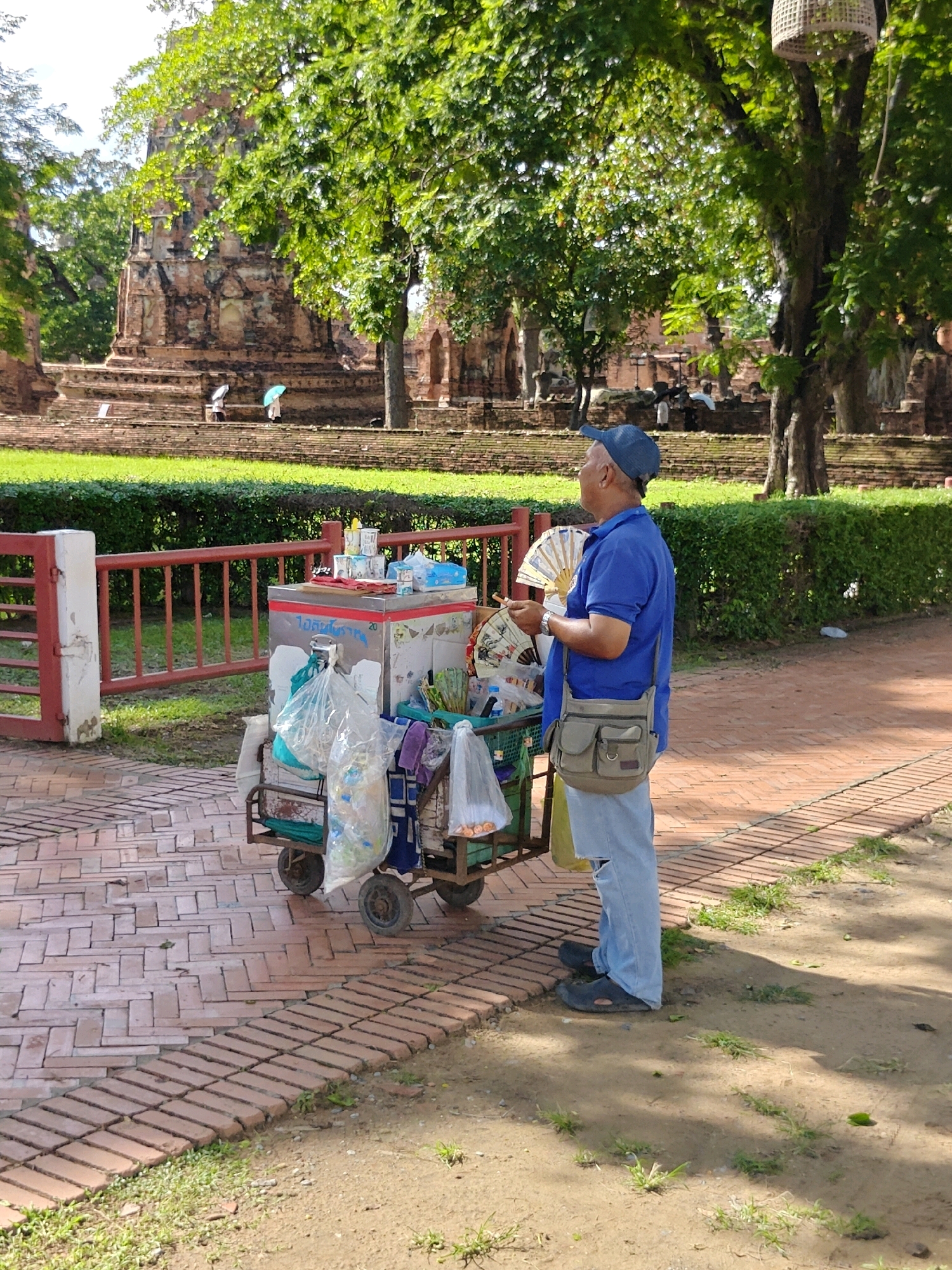 In Phra Nakhon Si Ayutthaya City Municipality, Thailand, a man wearing a blue shirt, blue cap, and light blue jeans stands next to a mobile cart filled with goods. The cart contains an ice chest, plastic bags with items inside, and various other products. The man holds a fan in his right hand and has a canvas bag slung over his left shoulder. The cart has two wheels on each side. Behind the man and the cart, a red metal fence separates the walkway from a grassy area. Further in the background, ancient brick ruins and lush green trees are visible. The ground is paved with reddish-brown bricks in a herringbone pattern. The overall scene suggests a vendor operating in a historical or tourist location.