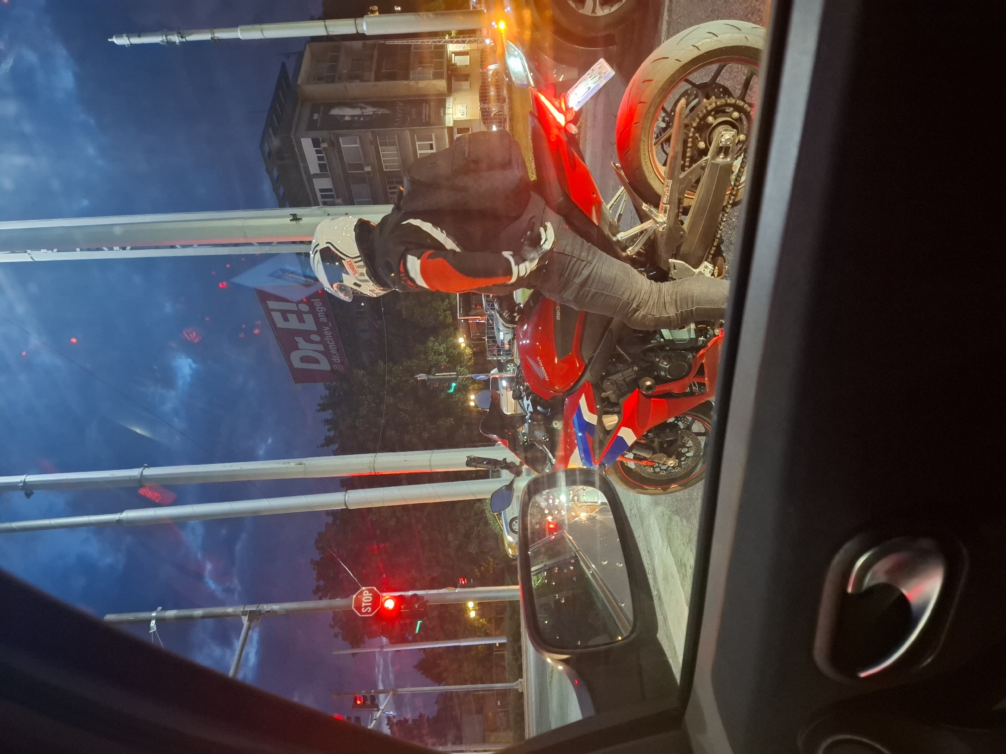 In the city of Unknown, Unknown, a motorcyclist wearing a white helmet with red and black accents, a black jacket with red and white stripes, and dark jeans is stopped at a traffic light, waiting for it to turn green. The motorcyclist is seated on a red and white Honda motorcycle. The image is taken from inside a car, showing the side-view mirror reflecting the traffic light and other vehicles. A "STOP" sign is visible on a traffic light pole in the background. The sky is dark blue with some clouds, indicating it's either dusk or dawn. Buildings and trees are visible behind the traffic light. The overall scene depicts a typical urban traffic situation at twilight.