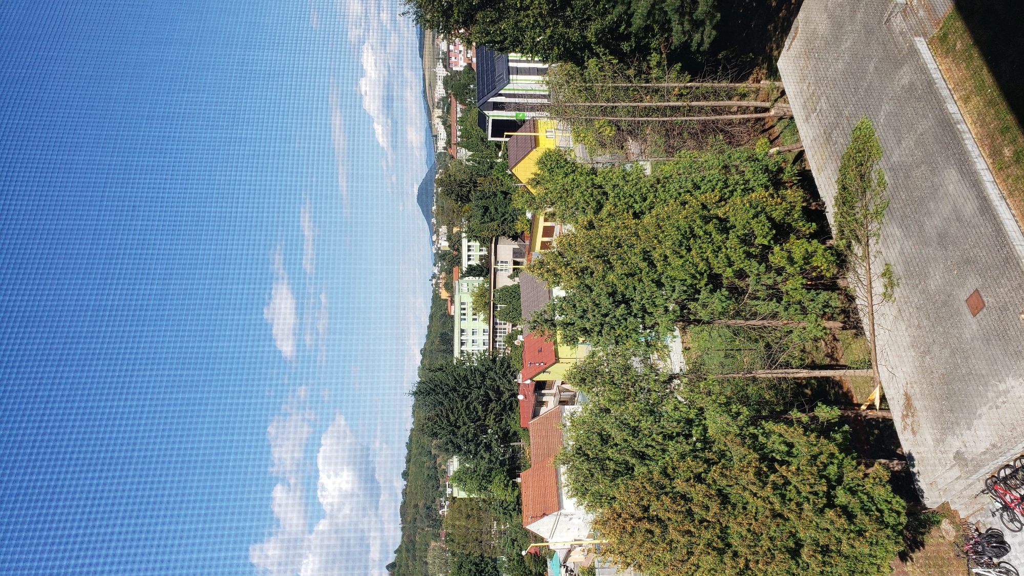 This image captures an aerial view of a residential area in Prešov, Slovakia, likely on a sunny day. In the foreground, a paved, multi-level surface with a grid pattern is visible, with several bicycles parked on the left side. A fallen tree branch lies across this surface. Behind this, a cluster of residential buildings with red and yellow roofs is nestled amongst lush green trees. Further in the background, larger apartment buildings, some with green and white facades, are visible, along with smaller houses. Rolling green hills form the backdrop to the city. The sky is a vibrant blue with scattered white clouds, and the entire scene is viewed through a mesh or screen.