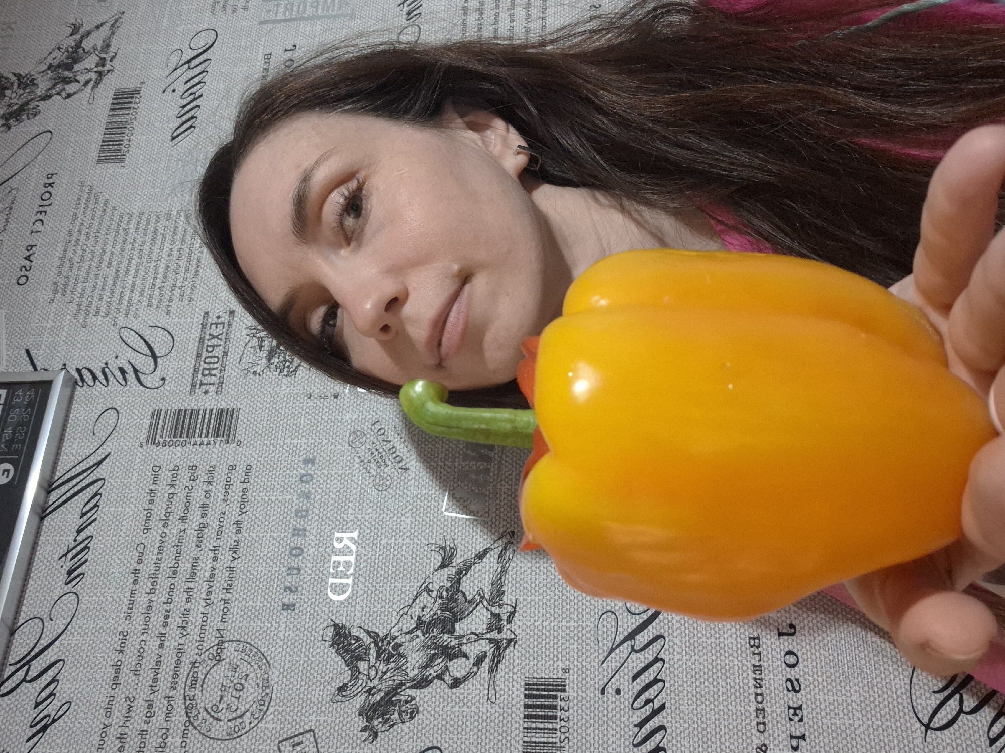 A person is lying down with their face near an orange bell pepper. The person appears to be holding the bell pepper in their hand. The background is a surface with text and drawings, including the word "RED" and barcode markings. The person has brown hair with a hint of pink or purple in it. The subject's hair is to the right of the subject's face. The scene appears to be an indoor setting. The location is unknown.