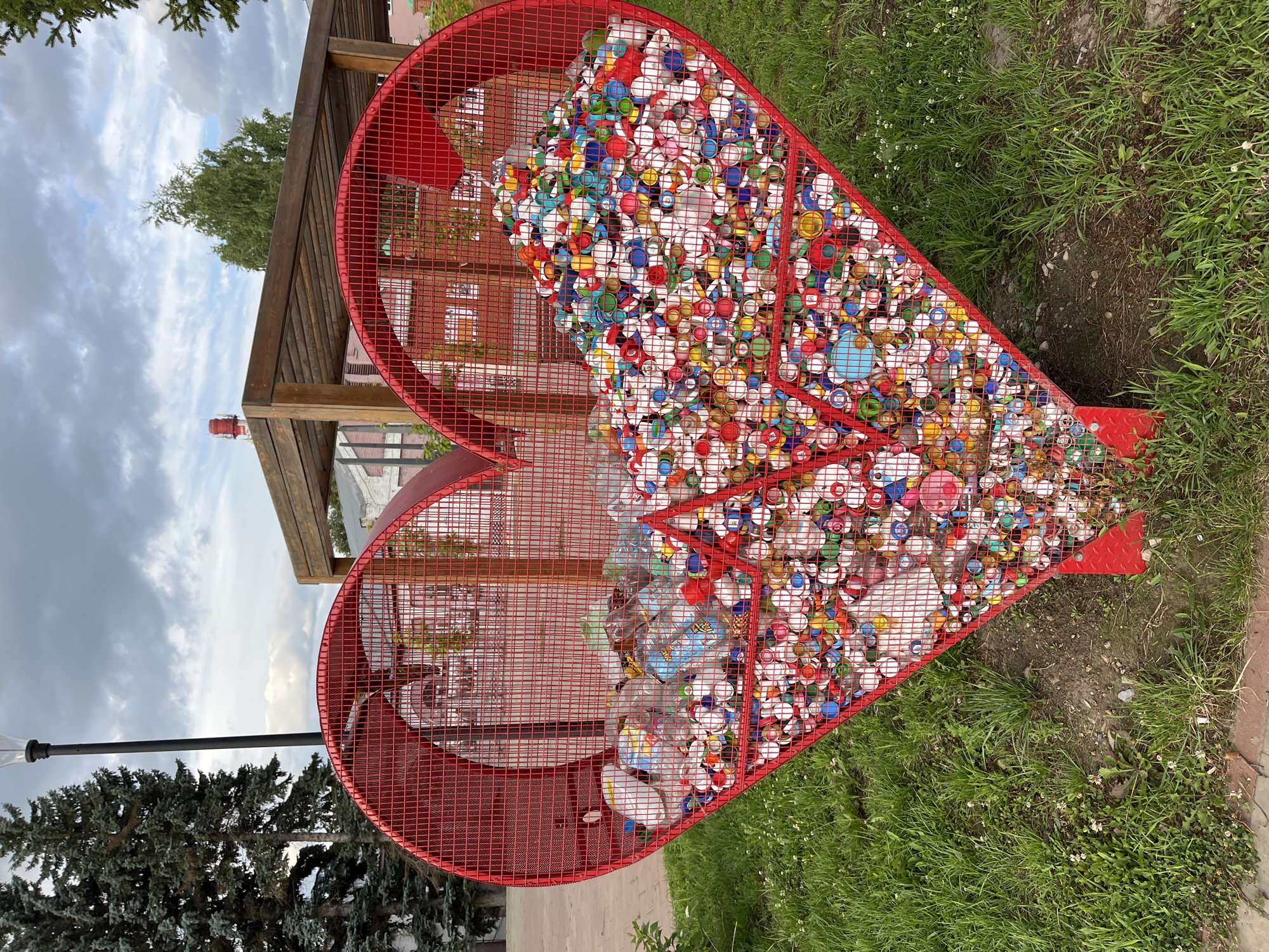 The image shows a large, red, heart-shaped metal bin filled with plastic bottle caps and some plastic bottles. The bin is located outdoors in what appears to be a park or public space with green grass and trees. In the background, there is a wooden structure, buildings, and a cloudy sky. The bin itself has a mesh construction and a red metal frame. The bottle caps are of various colors, including red, blue, green, yellow, and white, and they fill the majority of the heart-shaped bin. Some clear plastic bottles are also visible towards the top left of the bin. The scene is captured from a slightly low angle, looking up at the bin. The location is identified as Blagoveshchensk, Russia, though this specific detail cannot be verified from the image alone.