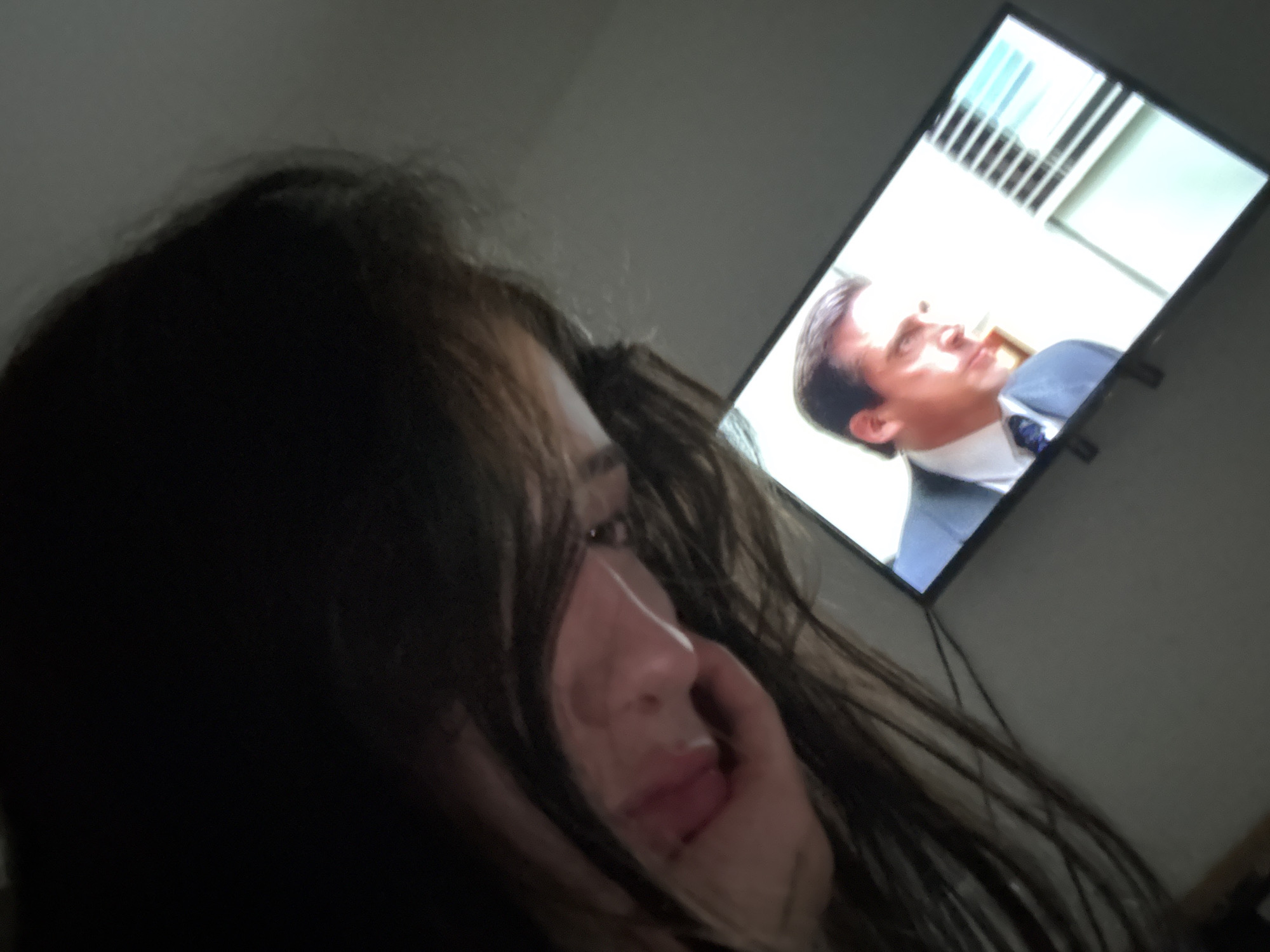 A woman with dark hair is watching television. On the screen is a scene from the American television show "The Office," featuring actor Steve Carell as the character Michael Scott. The room appears to be dimly lit. It is not possible to determine the location of this image in Buenos Aires, Argentina, as there are no features to indicate that this image was taken in that city.