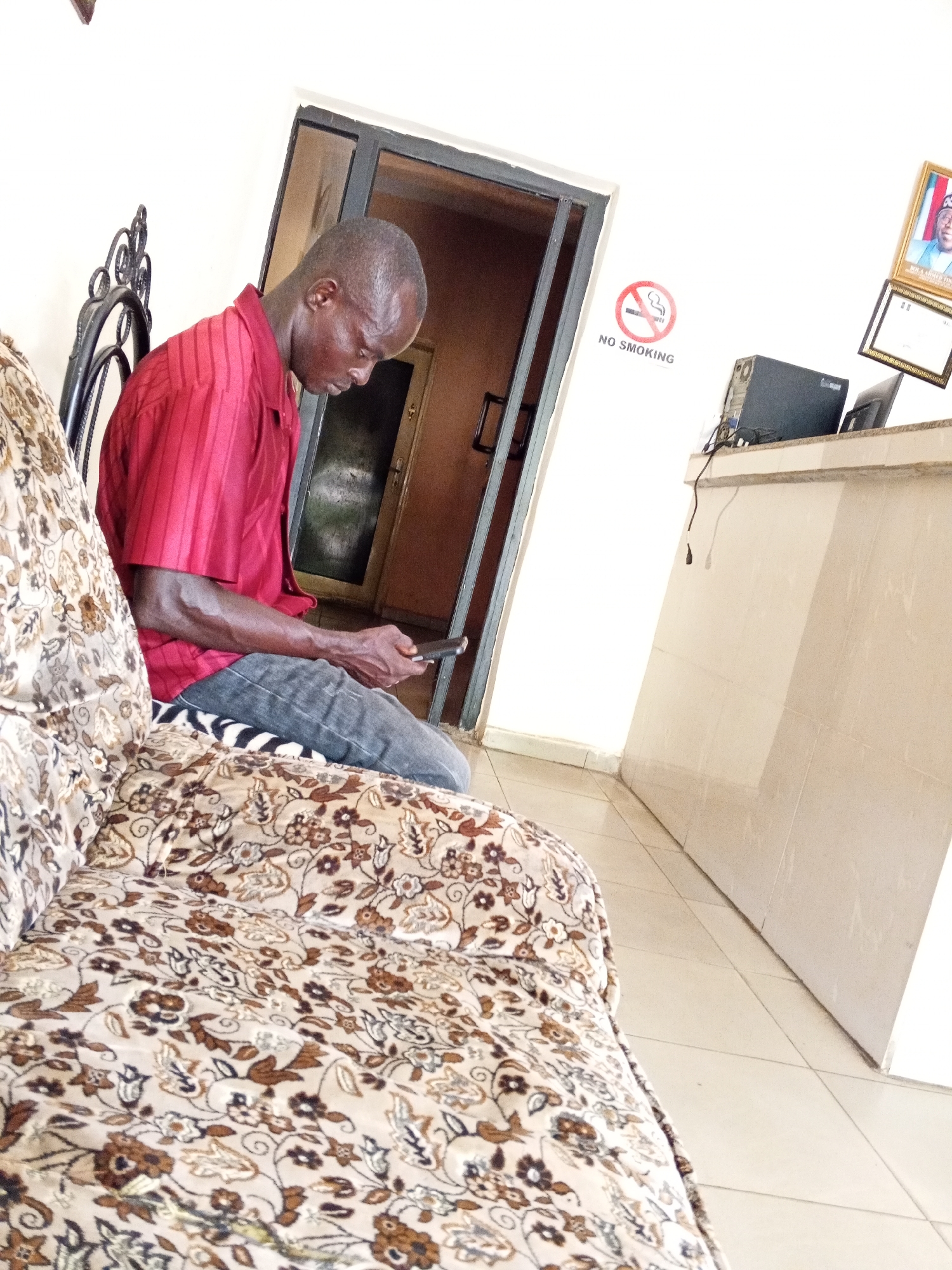 The image shows a man sitting on a patterned sofa, looking at his mobile phone. He is wearing a red shirt and grey jeans. The location appears to be inside a building, possibly a waiting area. A 'No Smoking' sign is visible on the wall, along with a framed picture and a computer setup on a counter. The city of location is unknown.
