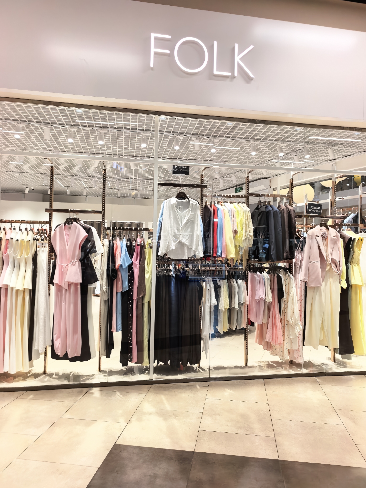 The image shows the exterior of a clothing store with the name "FOLK" displayed in large, white, illuminated letters on a white facade. The store has a large glass window displaying racks of women's clothing in various colors and styles, including dresses, blouses, and jackets. The clothing appears to be arranged by color and style, with a focus on pastel shades. Some of the garments have tags that read "НОВАЯ КОЛЛЕКЦИЯ" (New Collection) in Cyrillic script, indicating that the store is featuring its latest offerings. The interior of the store is visible through the window, showing a grid-like ceiling with track lighting. The floor in front of the store is tiled with large, light-colored square tiles, and sunlight is casting shadows on the floor. The image does not contain any recognizable landmarks or context that would definitively place it in Sterlitamak, Russia, or any other specific city.