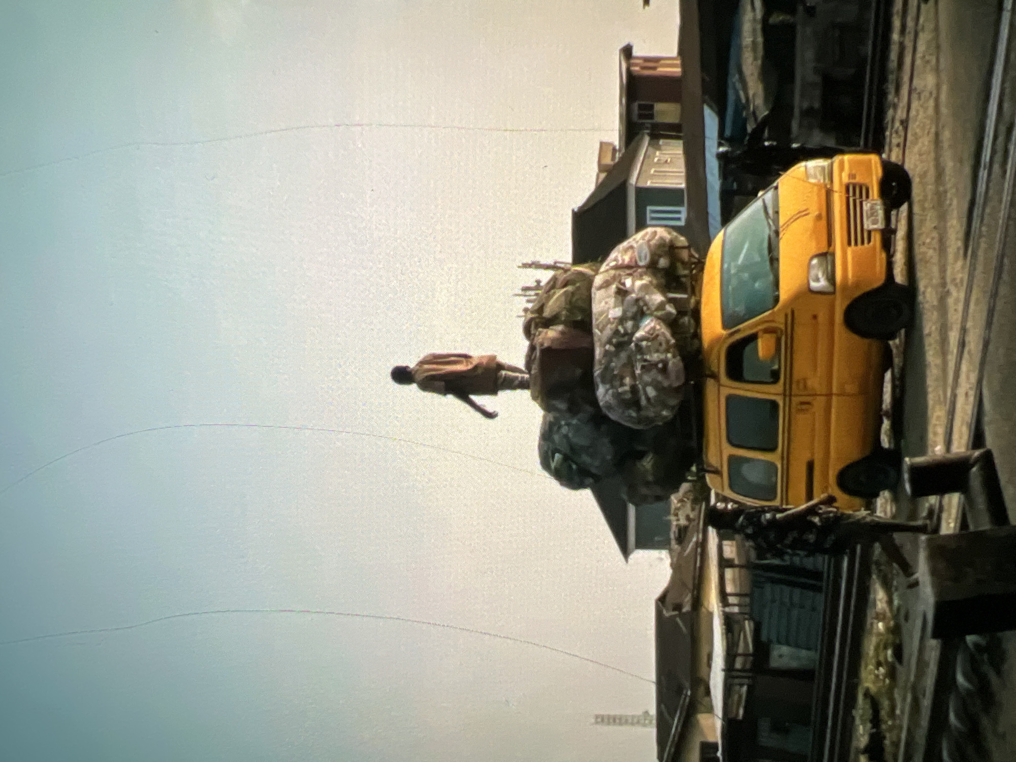A yellow minibus, overloaded with large, lumpy sacks of goods, is parked on a track, possibly a railway line, in an urban setting in Lagos, Nigeria. A young male figure stands atop the stacked cargo, appearing to balance precariously. In the foreground, a man in a patterned shirt walks from left to right, passing by the minibus. The background reveals a mix of low-rise buildings, some with corrugated metal roofs, and what appears to be market stalls or workshops. The sky is pale and hazy, suggesting a hot, possibly dusty, environment typical of Lagos. The scene captures a moment of transportation and commerce, highlighting the resourceful and often challenging methods of moving goods in the city.