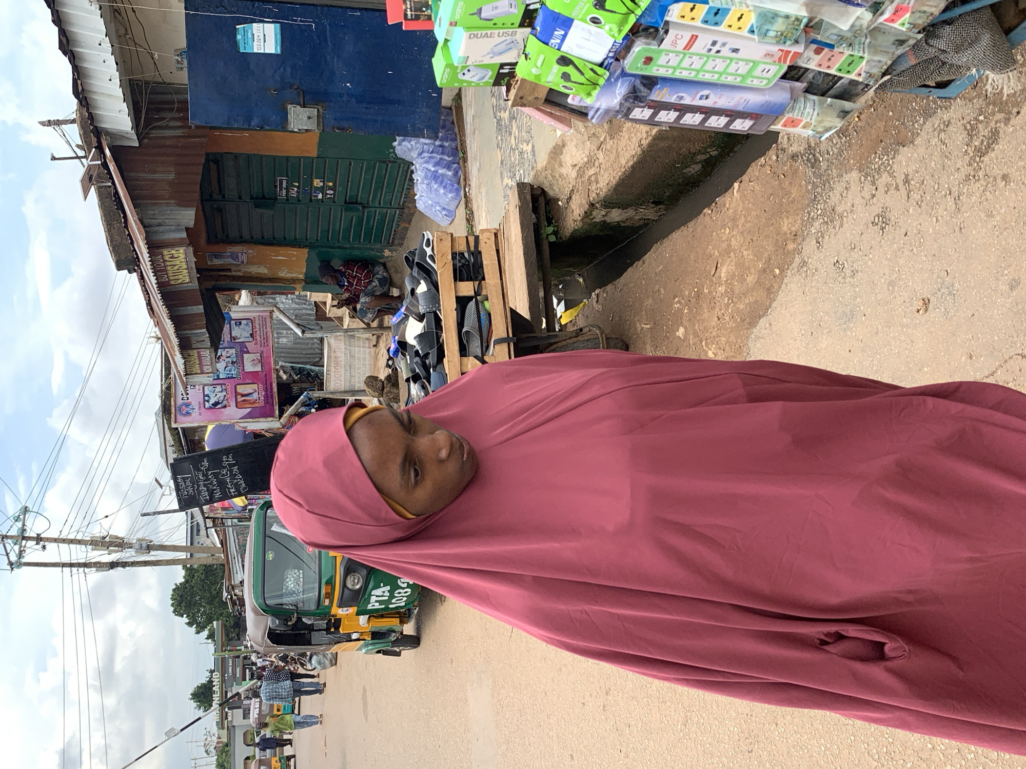 A young woman wearing a maroon hijab and a yellow under-scarf walks on a dirt road in Gura Top, Nigeria. She is facing away from the camera and looking down. Behind her, a yellow and green auto-rickshaw is parked on the side of the road. The auto-rickshaw has "PTA-1084" written on it. Several people, including men and women, are walking in the background.
To the right of the woman, there is a stall selling various electronic items, including phone chargers and power outlets. The stall is set up on the side of the road, with the items displayed on shelves and racks. Behind the stall, there is a building with a corrugated metal roof and blue doors. A man is sitting on a ledge in front of the building, and several bottles of water are stacked nearby. The environment appears to be a bustling market street with shops and stalls lining the road. The sky is cloudy.