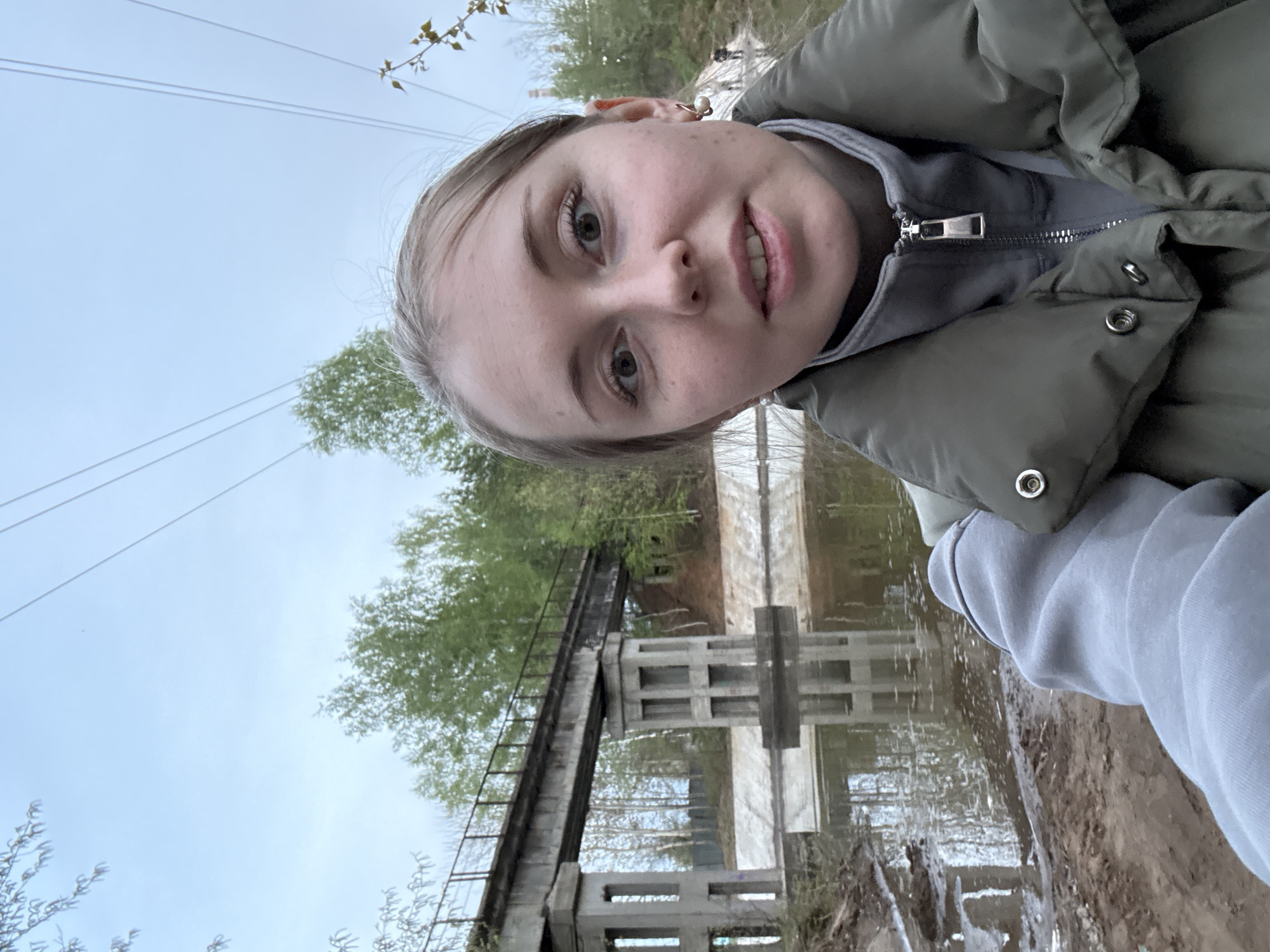 A selfie of a young woman with fair skin and blonde hair, wearing a green puffer vest and grey hooded sweatshirt. She is positioned in front of a concrete bridge structure that appears to be reflected in the water beneath it. Trees are visible behind the bridge, and power lines stretch across the sky. The ground appears muddy and wet. The location is Unknown, Unknown.