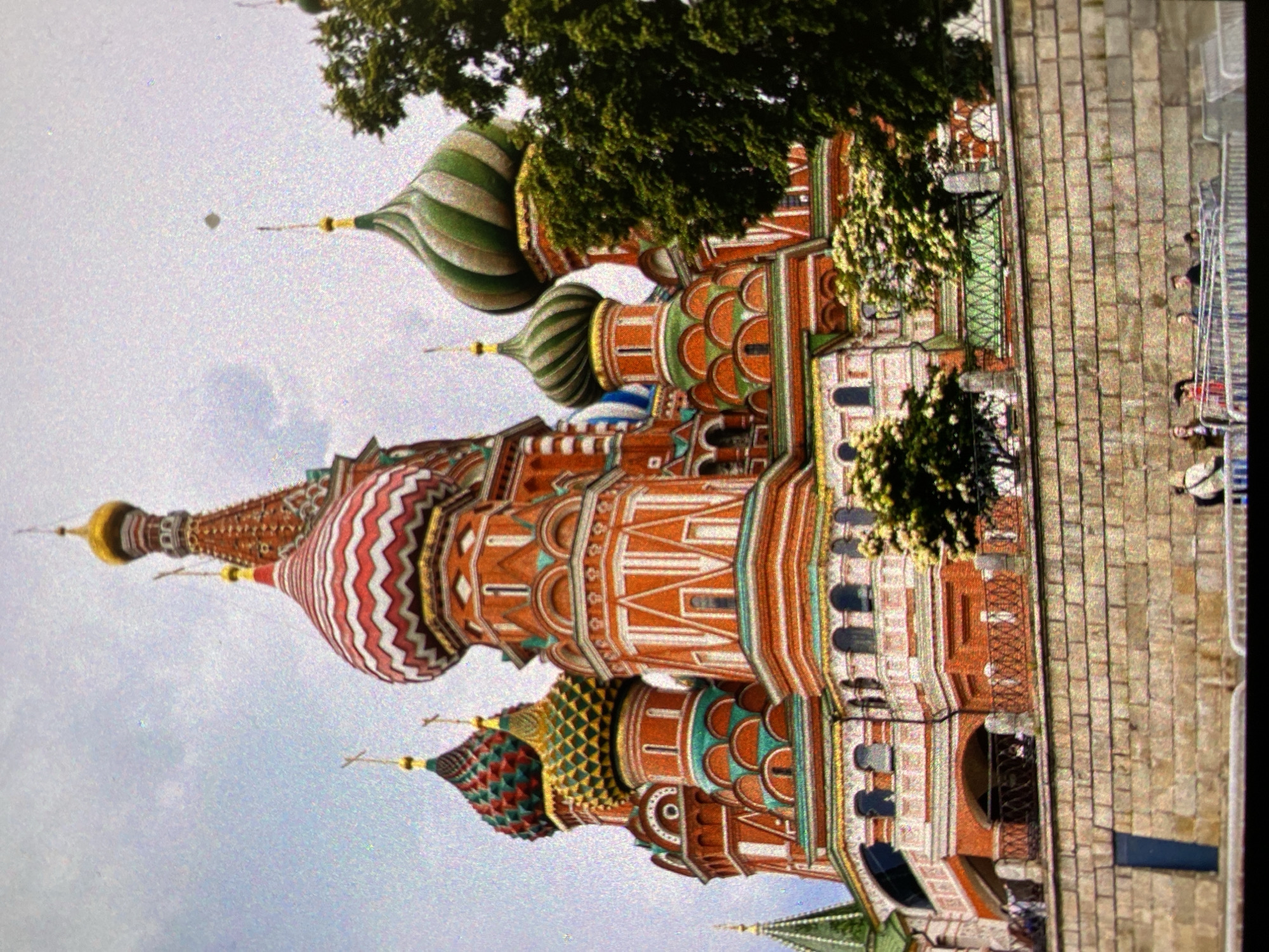 The image depicts Saint Basil's Cathedral in Moscow, Russia. The cathedral is a multi-domed church with colorful and patterned onion domes. The architecture features red brick walls with white and colored decorative elements. A stone pavement or walkway is visible next to the cathedral, with a metal fence. People are seen walking along the walkway. Trees are present, partially obscuring some sections of the building. The sky is visible behind the cathedral.