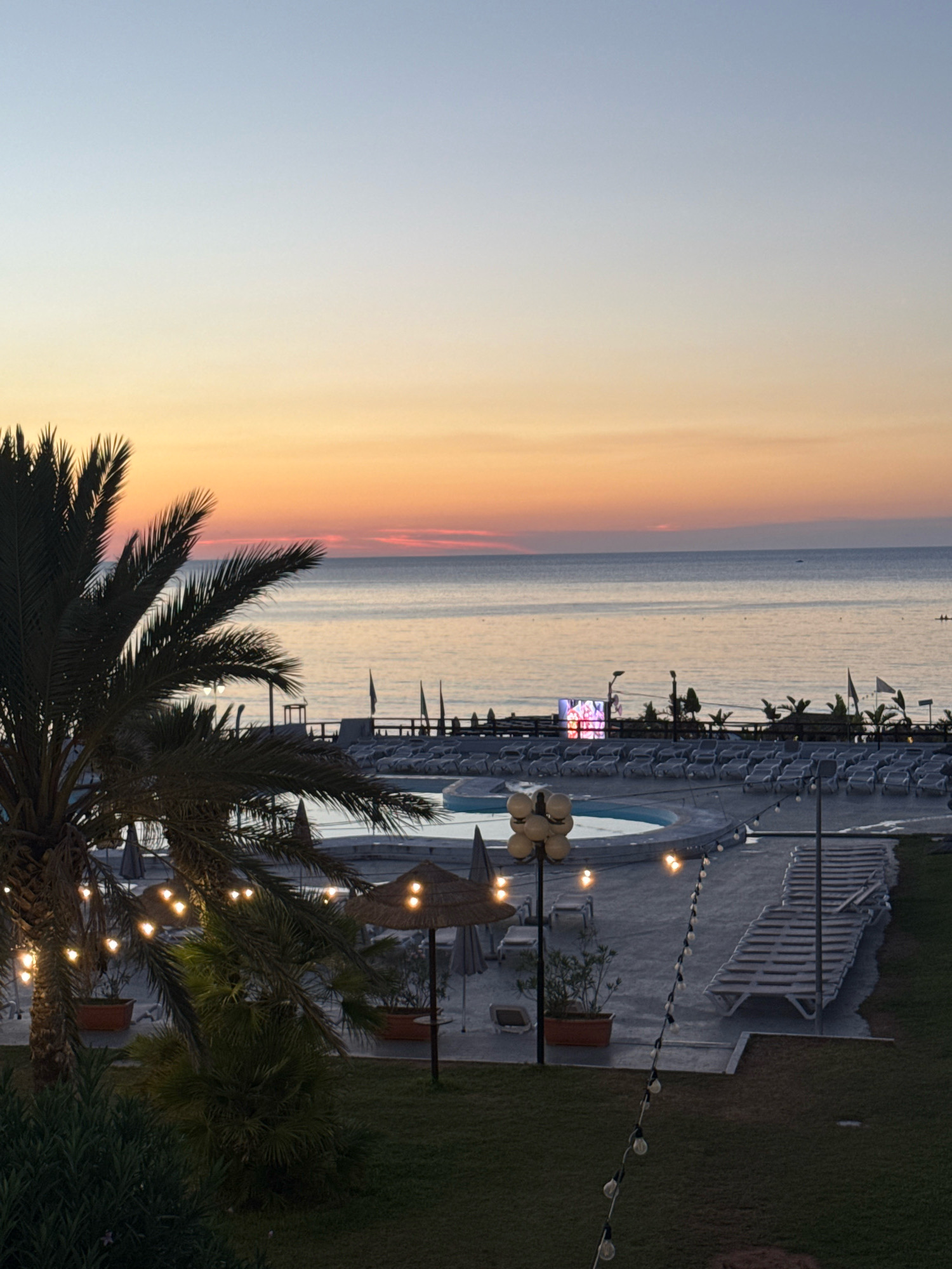 The image depicts a seaside resort at dusk in Isola delle Femmine, Italy. A large palm tree dominates the left foreground, its fronds silhouetted against the soft glow of string lights. In the mid-ground, a swimming pool is visible, surrounded by rows of empty lounge chairs. Beyond the pool area, the sea stretches out to the horizon, reflecting the warm hues of the setting sun. A large screen displays colorful graphics on the far side of the pool. The overall scene conveys a sense of tranquility and impending evening relaxation.