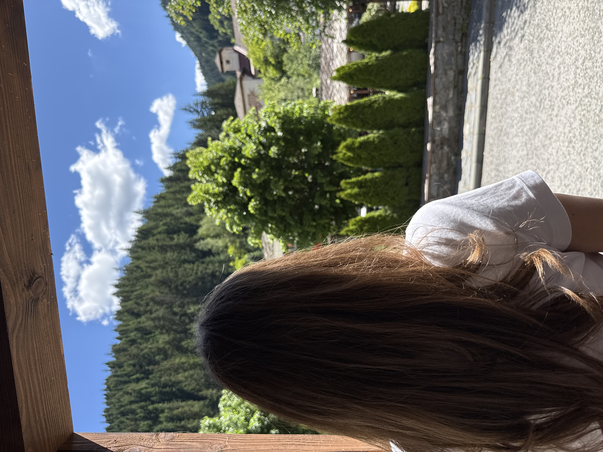 A young person with long, brown hair, viewed from behind, stands looking out at a scenic view of a mountainside covered in dense green pine trees under a bright blue sky with scattered white clouds. In the mid-ground, several conical evergreen trees are planted in a row, backed by a stone wall and some traditional Bulgarian houses with red tile roofs, one featuring a prominent tower. The scene is framed by a wooden overhang in the foreground, suggesting the person is looking out from a covered porch or balcony. The overall impression is one of peaceful contemplation in a rural mountain village setting, likely Shiroka Laka, Bulgaria.