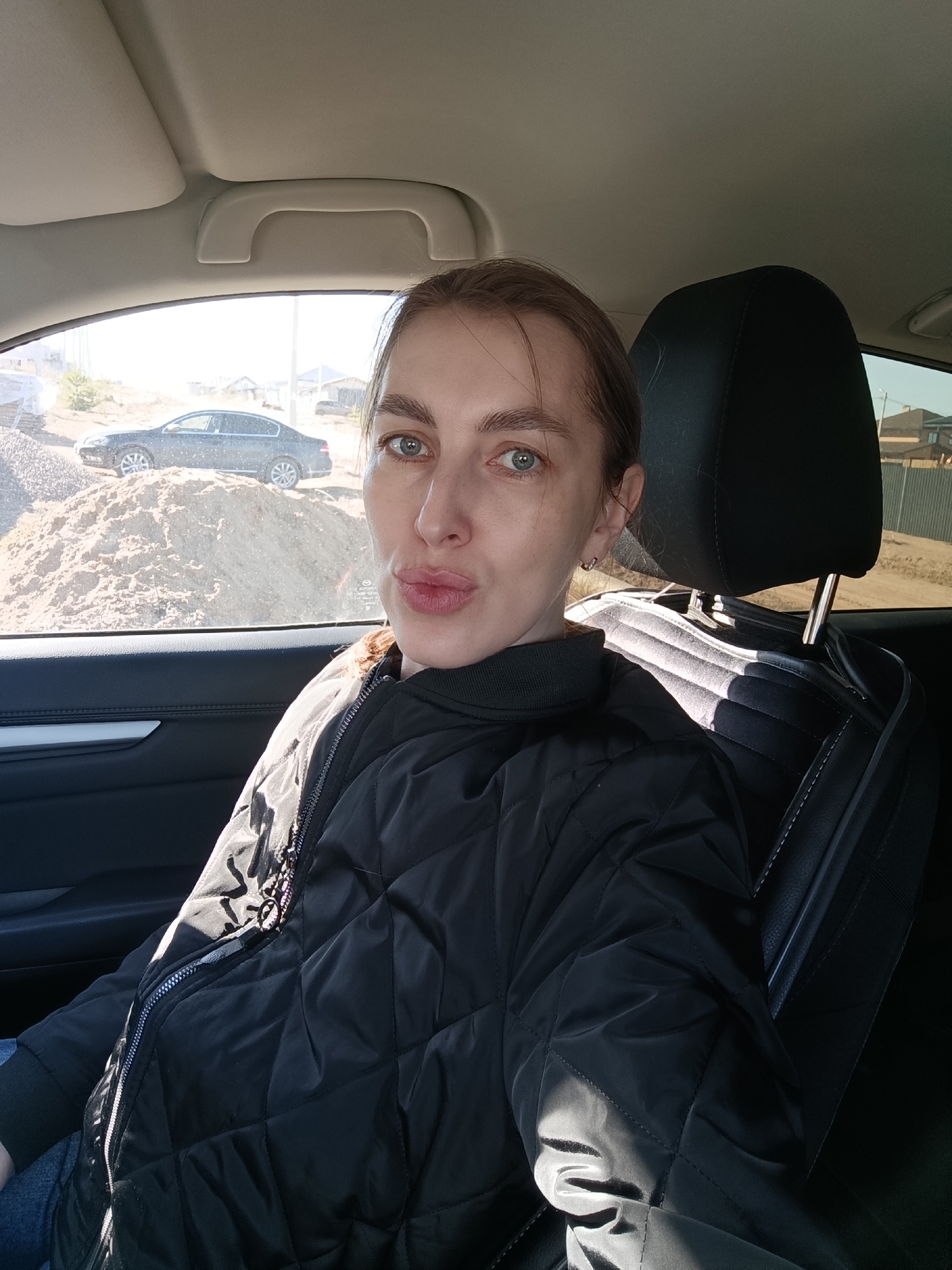 A woman sits in the driver's seat of a car. She wears a black quilted jacket. Outside the window, a mound of sand and gravel is visible, with a black sedan parked atop it. A house is also visible in the background. The scene is possibly located in the city of Chaikovsky, Russia.