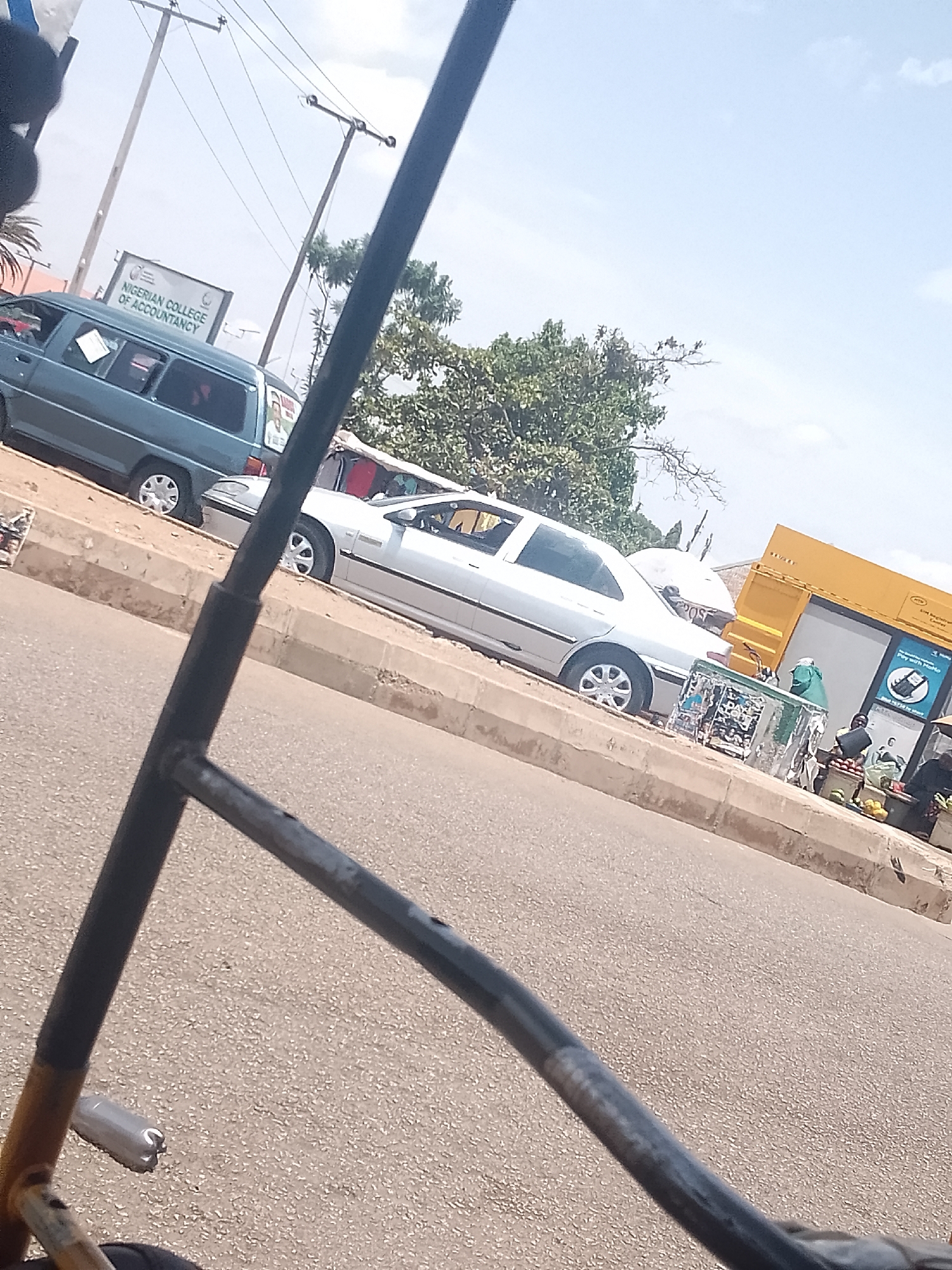 The image captures a street scene in Bukuru, Nigeria, observed from within a vehicle (likely a tricycle or similar). The focus is on the street, featuring a blue van parked adjacent to a sign reading "NIGERIAN COLLEGE OF ACCOUNTANCY". A silver car is parked next to the van. On the right side of the frame, a yellow booth or structure can be seen, along with what appears to be a vendor stall and people. The road surface is visible and occupies a significant portion of the lower half of the image. The sky is partly visible in the background. The perspective is slightly skewed due to the angle of the shot.