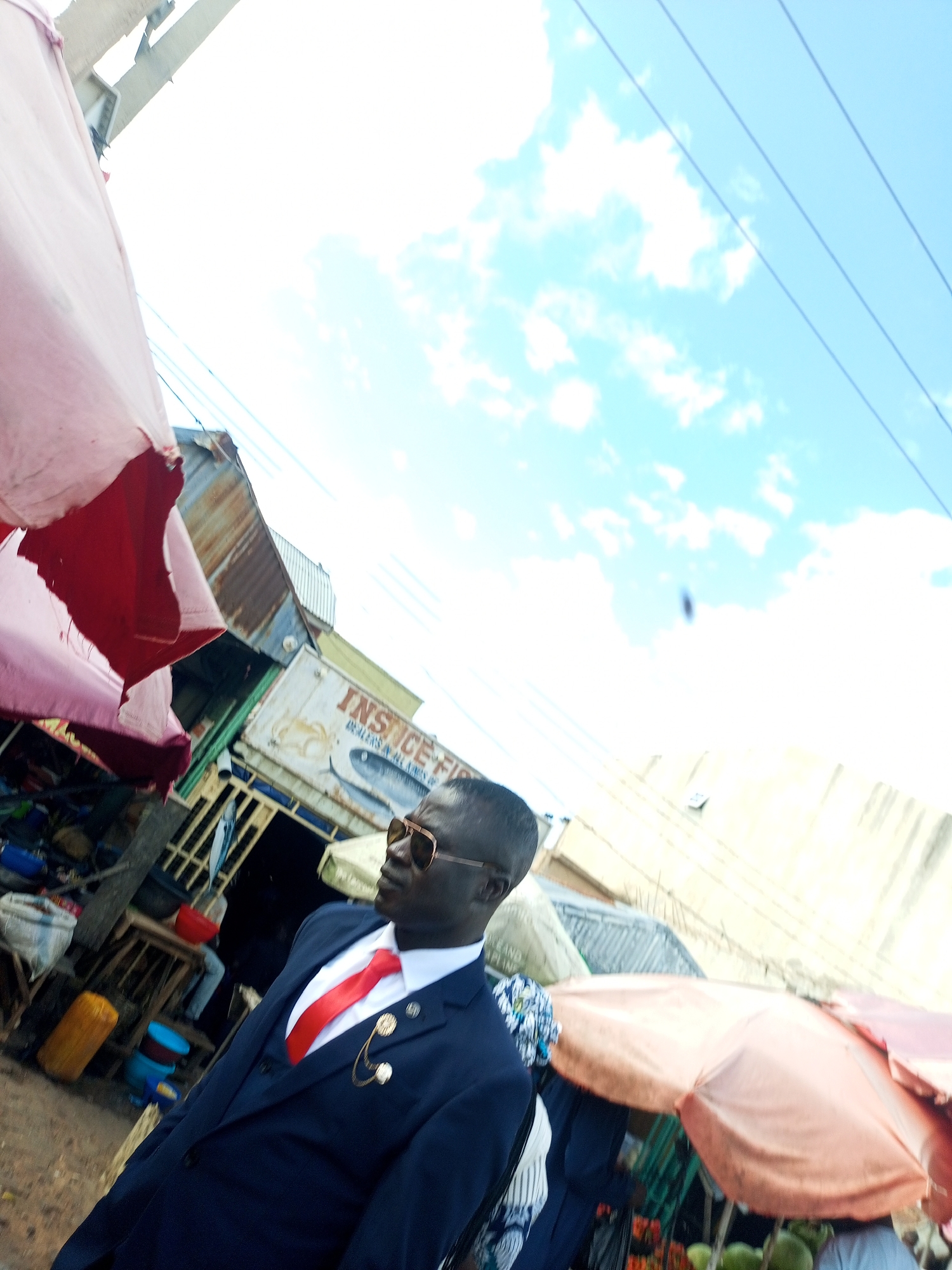 The image shows a man wearing a navy blue suit, white shirt, and red tie, with gold-rimmed sunglasses. He is standing in what appears to be an outdoor market, with stalls and vendors in the background. The market setting includes various goods, umbrellas, and corrugated metal structures. The sky above is blue with scattered clouds, and power lines are visible. The man is looking to his right, with his profile partially visible. His suit is adorned with a gold chain and two decorative pins on the lapel. The overall scene suggests a busy marketplace in Tempure, Nigeria.