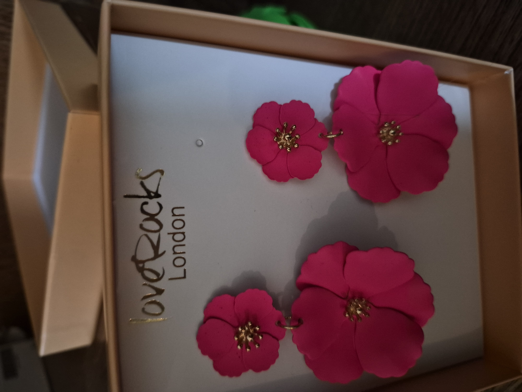 A close-up, eye-level shot captures a pair of bright pink floral dangle earrings presented in a light brown, open box. The earrings are the primary focus, with each featuring two flower-shaped drops. The upper flower is smaller and appears to be attached directly to the box's backing, while the lower, larger flower hangs below it. Both flowers have a delicate, scalloped edge to their petals and a gold-colored center with protruding stamens. Above the earrings, the brand name "love Rocks London" is displayed in a stylized gold font on the box's white interior. The box itself is slightly angled, revealing the inside and a portion of its outer structure. The background is softly blurred, with hints of dark wood and a touch of green. The lighting creates subtle shadows on the earrings and the box.