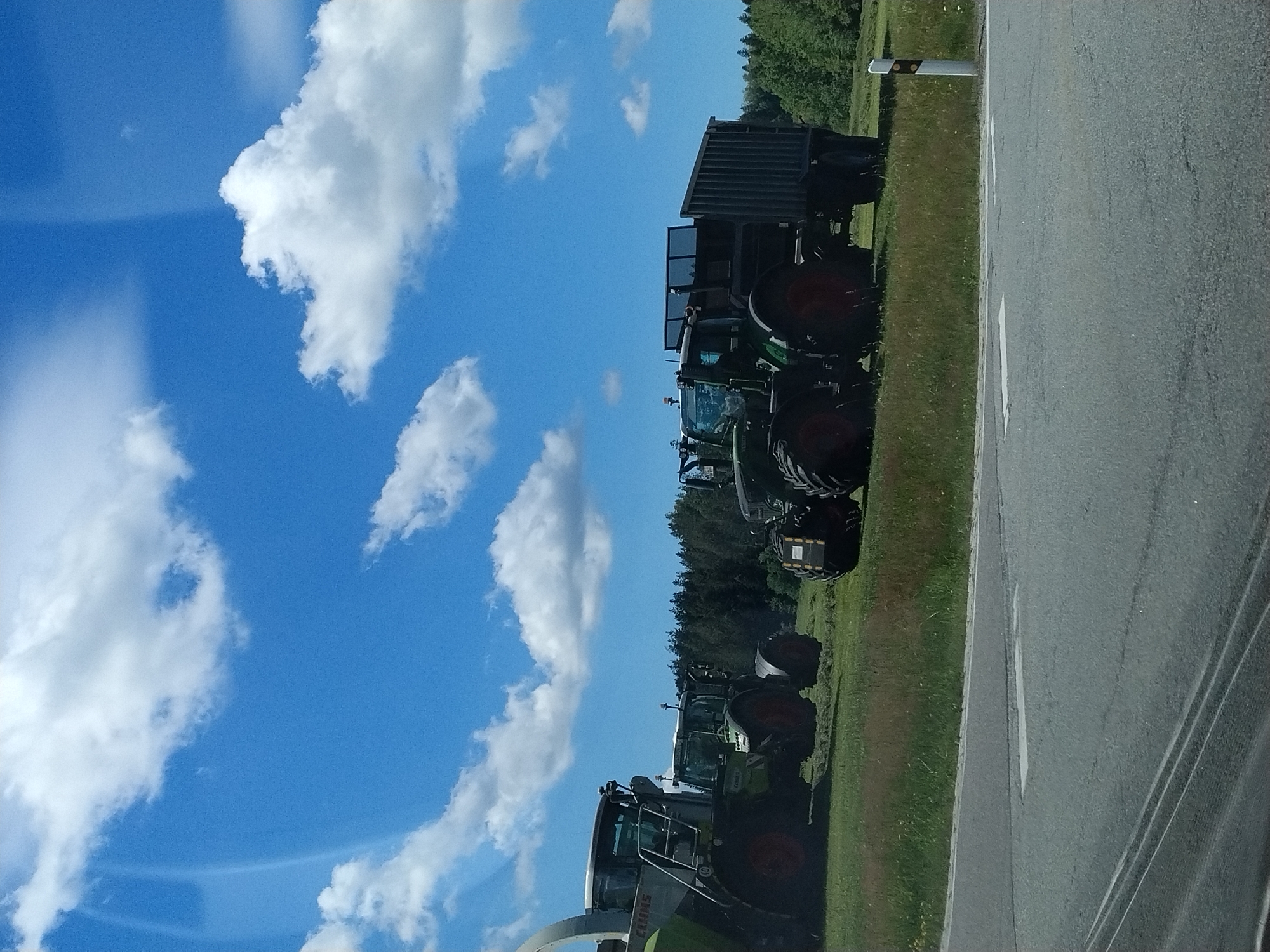 The image depicts an outdoor scene on a sunny day, likely in a rural area near Hinterschmiding, Germany. In the foreground, a grey asphalt road is visible with white lane markings. Beyond the road, a grassy field stretches out, dotted with some wildflowers. Several agricultural machines are parked on the field.
On the left side of the image, a large, green and grey CLAAS forage harvester is positioned. To its right, two green Fendt tractors are parked. The tractor on the left is smaller than the one on the right. The Fendt tractor on the right is connected to a dark grey, large-capacity trailer. In the background, a dense line of dark green trees forms a forest. The sky is bright blue with scattered white, fluffy clouds, suggesting fair weather. No people are visible in the frame. The overall impression is one of agricultural machinery at rest, likely during a break in field work.