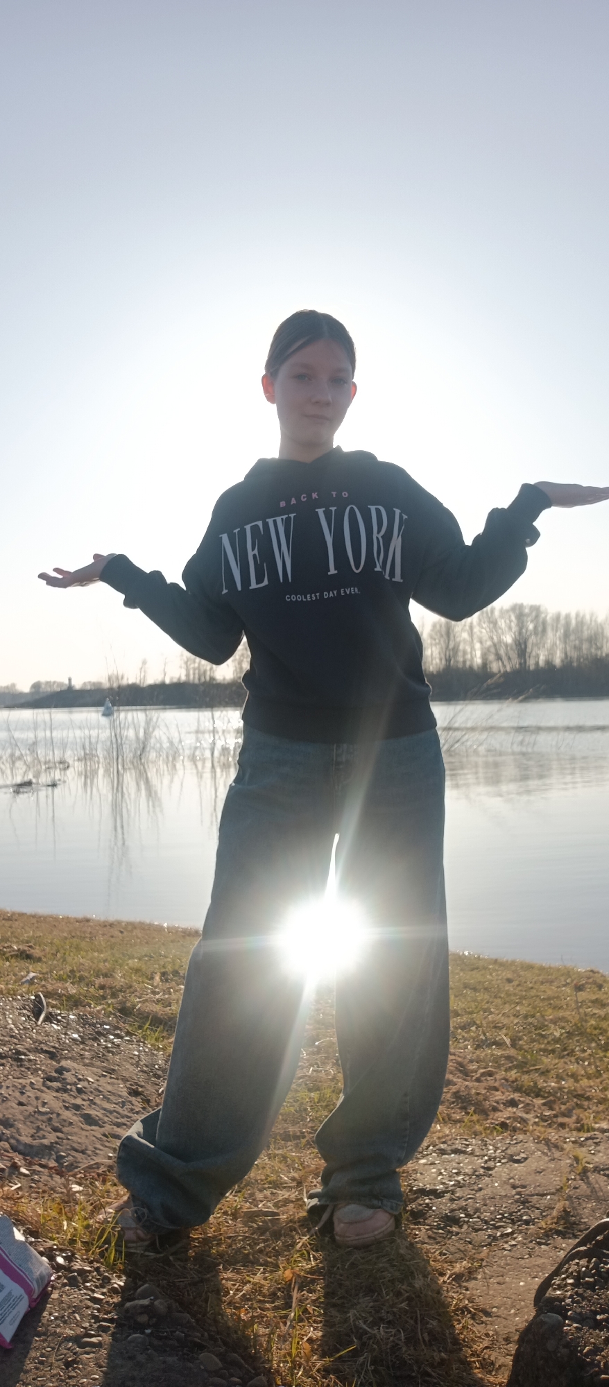 A young person stands on a dirt and grassy area near a body of water. They are wearing a dark hoodie with the words "BACK TO NEW YORK, COOLEST DAY EVER" printed on it, along with loose-fitting blue jeans and sneakers. The person has their arms outstretched. The sun is bright, causing glare in the lower half of the image. In the background, there is a body of water with sparse vegetation along the opposite shore. The location is identified as Unknown, Unknown.