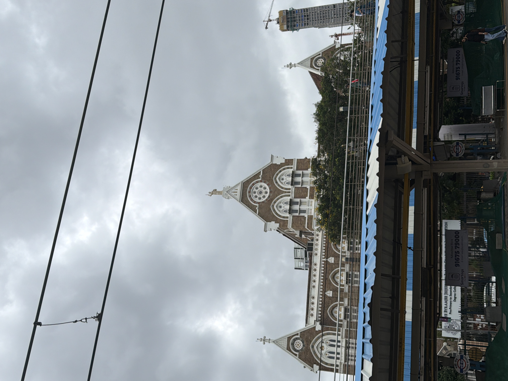 This is a photograph taken on a cloudy day, showing the facade of a historic brick building with arched windows and decorative white trim, likely a public building or institution, in Mumbai, India. A statue stands atop one of the building's pointed gables. In the foreground, a modern train station platform with a blue and white corrugated roof is visible, with overhead wires for electric trains stretching across the sky. To the right of the historic building, a tall, modern skyscraper under construction is partially visible, with a crane on top. People are seen on the elevated walkway of the train station, and at ground level near the platform. A banner advertising a school is attached to a fence in the lower part of the image. The overall atmosphere is overcast and typical of a monsoon season day in Mumbai.