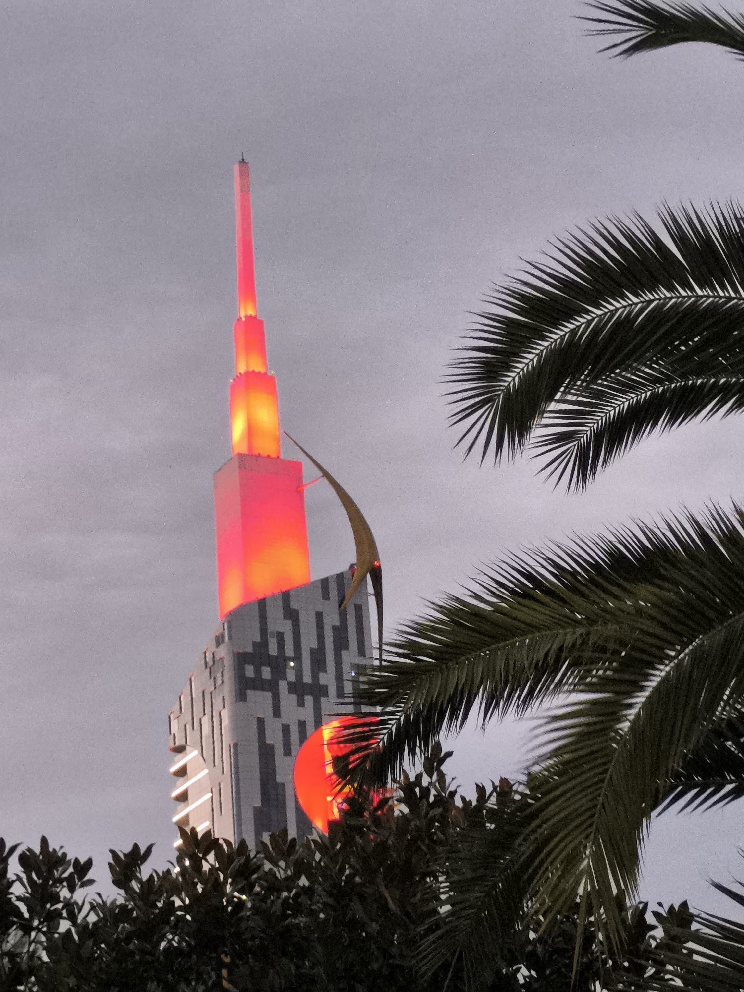 The image shows the Hilton Batumi, a skyscraper in Batumi, Georgia, illuminated with red and orange lights against a cloudy sky. The building features a distinctive spire that tapers to a point, and a curved golden architectural element is visible on its side. In the foreground, palm fronds and dark green foliage partially obscure the lower portion of the skyscraper and a large, red, circular illuminated object. The overall scene suggests an evening or twilight setting.