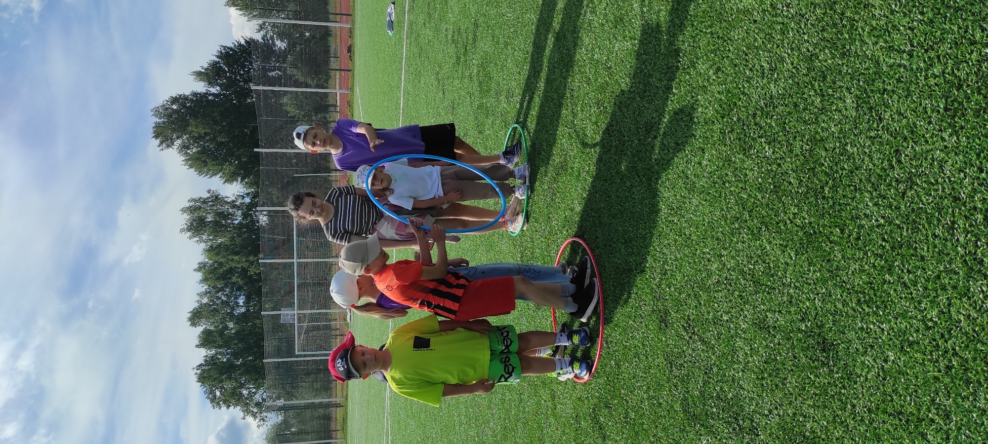 A group of children and two adults are gathered on a bright green artificial turf field under a partly cloudy sky. In the foreground, a boy in a neon green t-shirt and black shorts with "Respect" written on them stands inside a pink hula hoop. Next to him, another boy in an orange and black striped shirt and red shorts is inside a pink hula hoop with a blue hula hoop around his waist, held by another child. A girl in jeans and a white t-shirt with a blue hula hoop around her is also present. To the right, a boy in a purple t-shirt and black shorts stands near a green hula hoop. Behind the children, an adult woman with dark hair wears a striped t-shirt and a plaid shirt draped around her shoulders. Another adult, a young woman with glasses, stands in the background wearing a striped t-shirt. The field appears to be a sports field, possibly a soccer field, with a fence and trees visible in the background. The location is identified as Noviy, Russia.