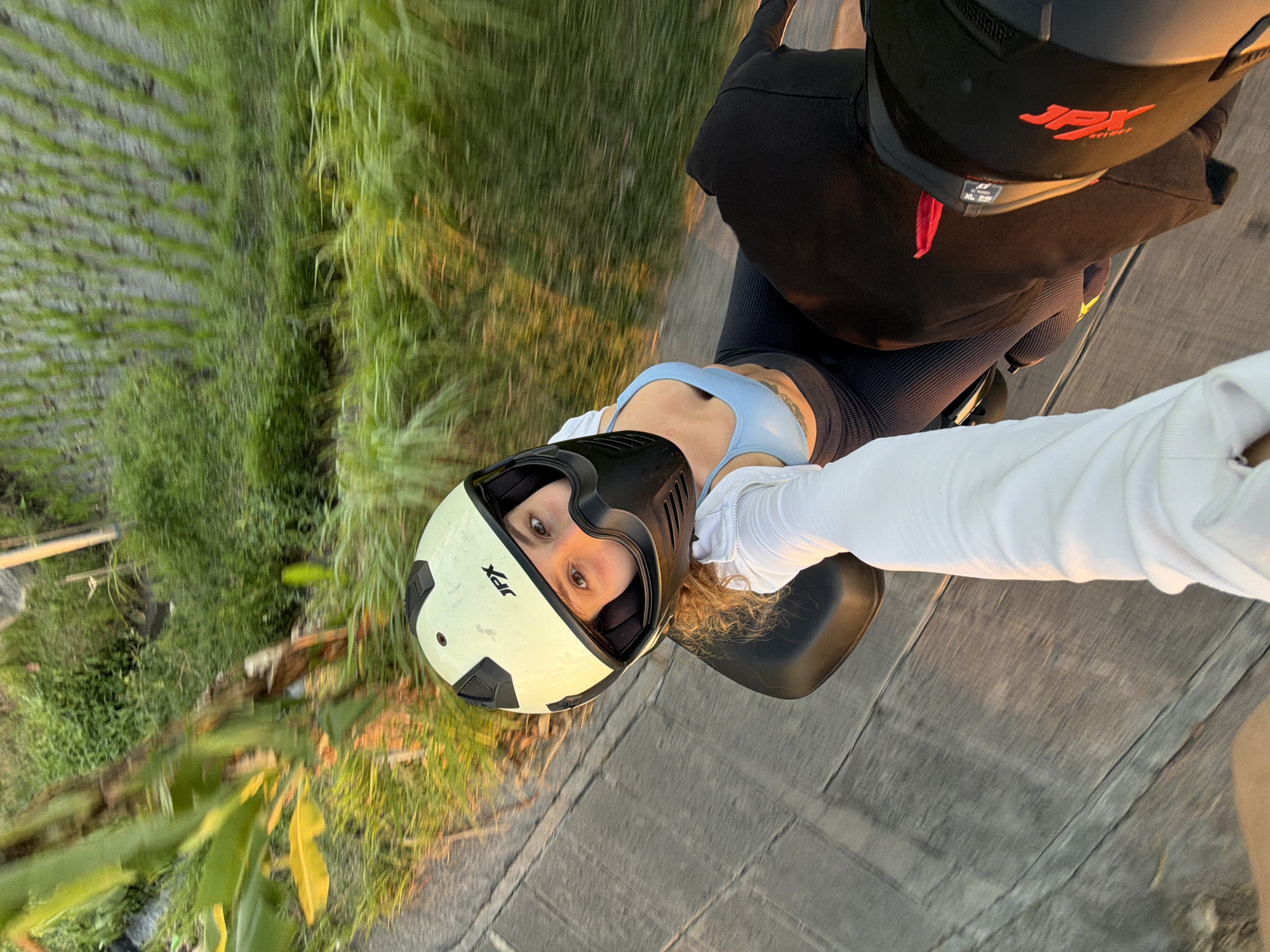 A first-person perspective shot captures a woman wearing a light yellow helmet with black accents and the logo "JPX" on it. She is on the back of a motorcycle, wearing a light blue sports bra and dark leggings. Her right hand, gloved in white, is extended towards the camera. The motorcycle is on a paved path with gray wooden planks. To the right, lush green vegetation is visible, suggesting a rural or tropical environment, possibly rice paddies as seen in the blurred background. The setting appears to be the outskirts of Mengwi, Indonesia, given the common landscape of rice fields and the warm, natural lighting indicative of an outdoor setting during a golden hour.