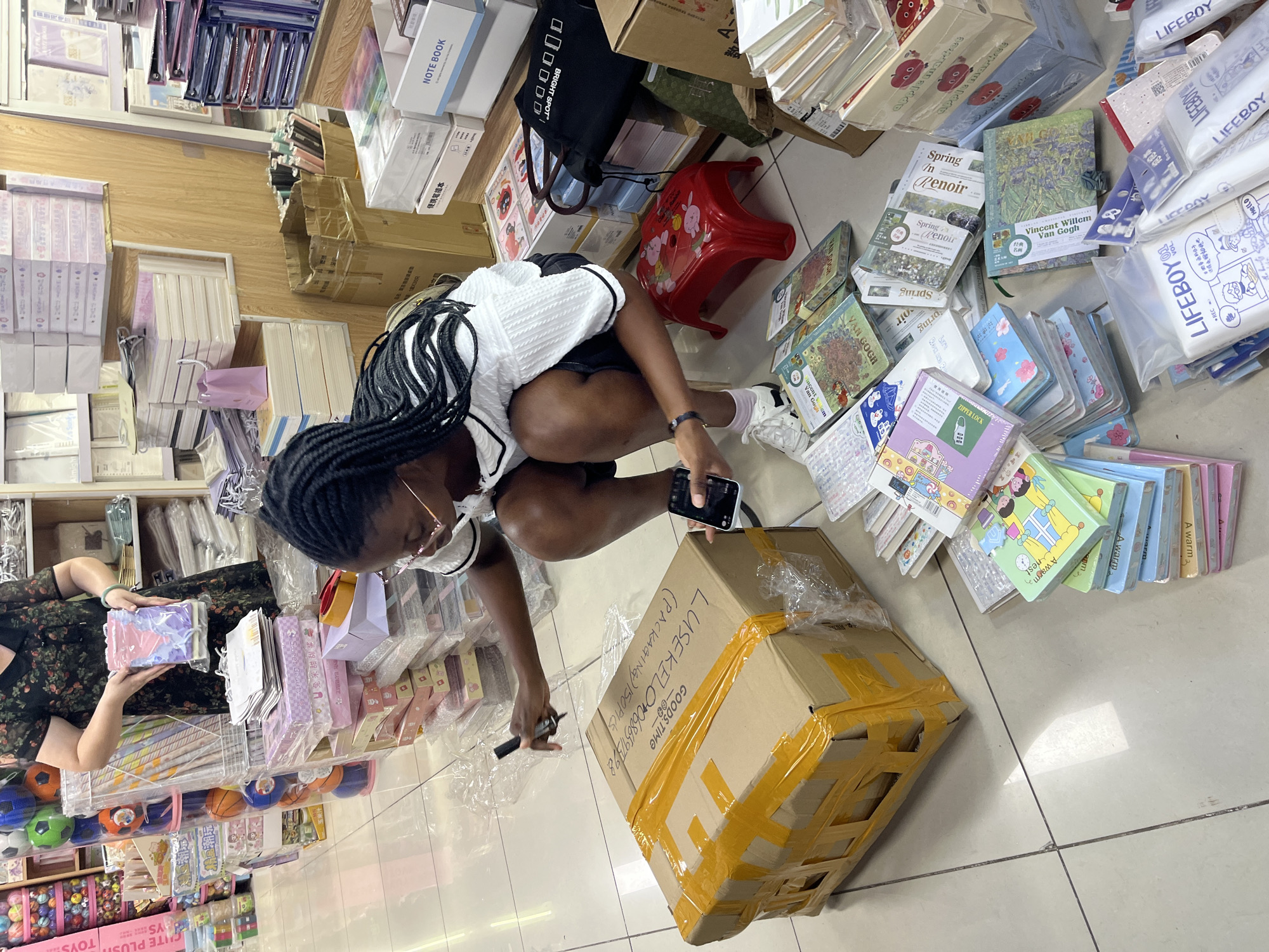 In Liwan District, China, a young Black woman wearing glasses and a white top with black trim is squatting next to a cardboard box. She holds a black marker in her right hand and is poised to write on the box. Her left hand holds a smartphone, and she appears to be scanning a barcode with it. The box is taped with yellow packing tape. She is surrounded by stacks of stationery, notebooks, and books on a tiled floor. In the background, shelves are filled with more stationery items, and another person is visible in the upper left corner of the image.