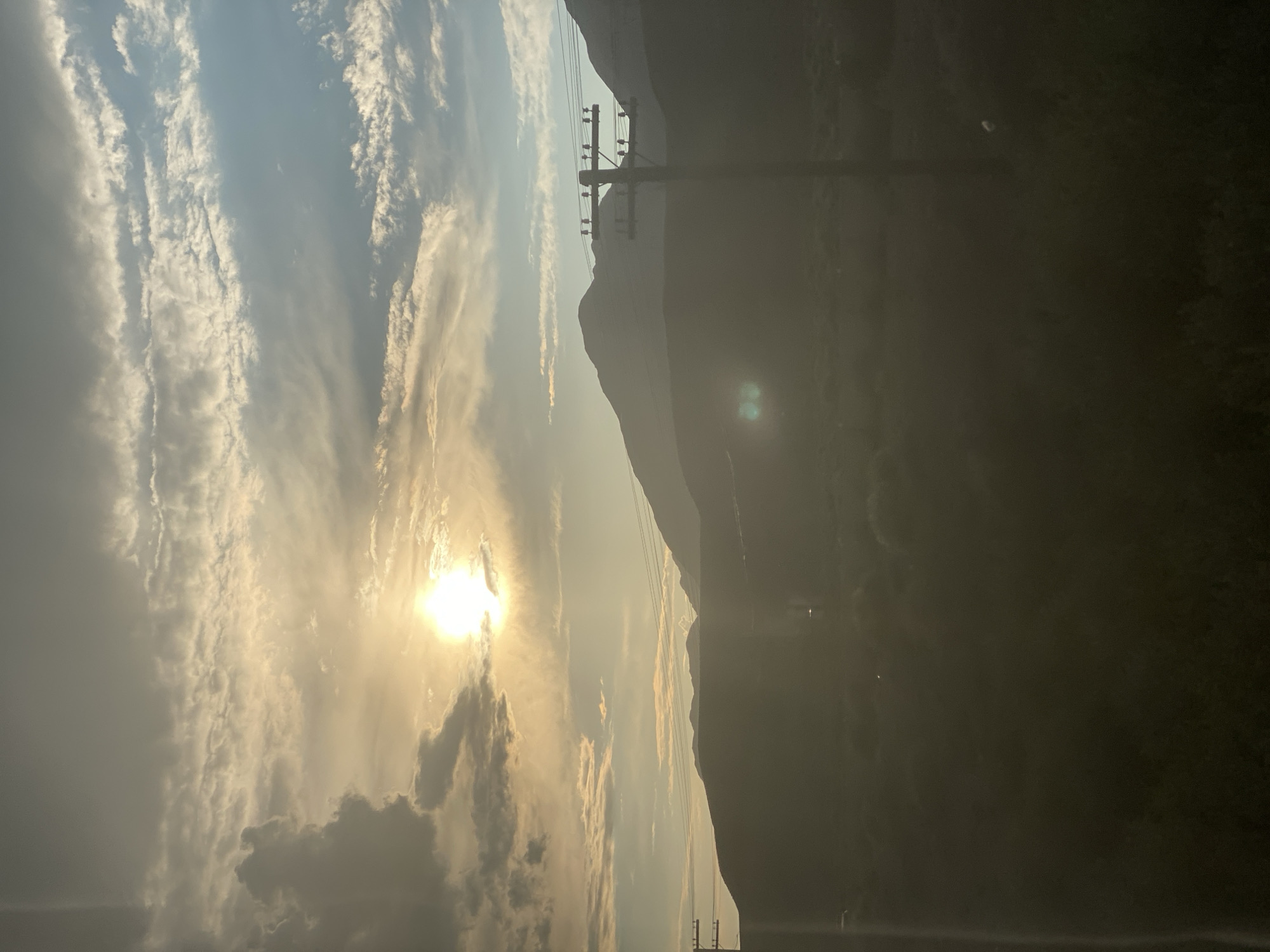 The image depicts a mountainous landscape at sunset or sunrise, with the sun partially obscured by clouds. The sky is a blend of blue and hazy yellow tones. In the foreground, there is a silhouette of power lines supported by a utility pole. The mountains are dark and shadowy, with a soft glow emanating from the horizon. The lower portion of the image shows a dark, out-of-focus landscape, possibly fields or low vegetation. There are no people or animals visible in the scene. The location is identified as Kok-Moinok - 1, Kyrgyzstan.