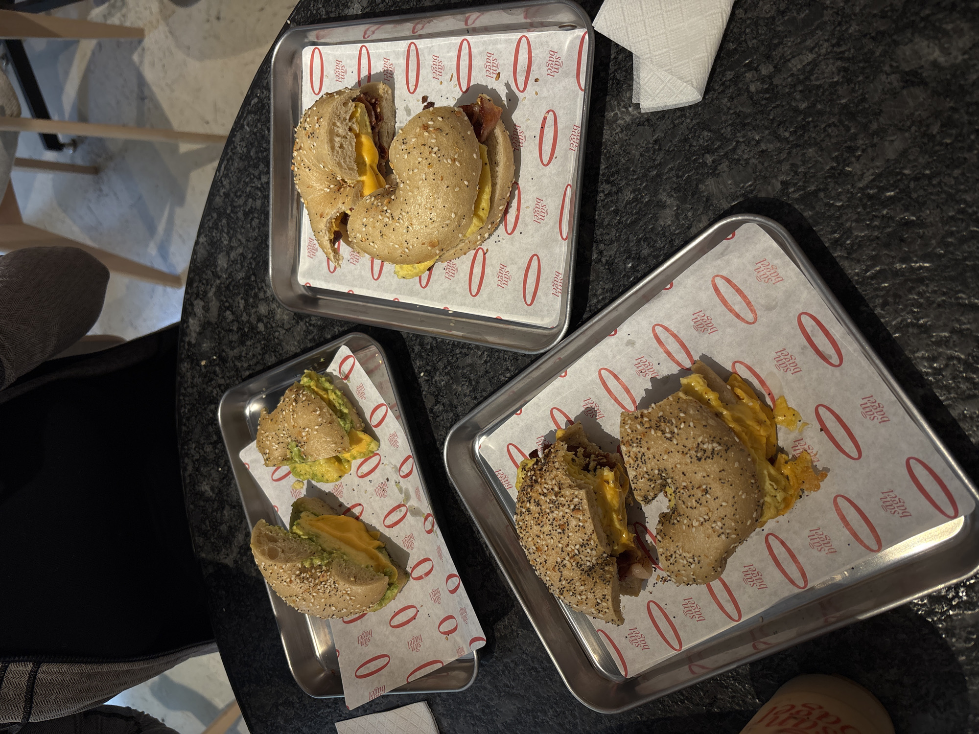 The image shows three small metal trays, each containing a bagel cut in half and filled with various ingredients. The trays are placed on a dark, patterned surface, possibly a table or counter. One tray holds a bagel with egg and bacon, another contains a bagel with egg and avocado, and the third has a bagel filled with egg and cheese. A crumpled white napkin is visible near the top-right tray. The trays are lined with paper that has "San Bagel" written on it. The image appears to be taken from an overhead perspective.