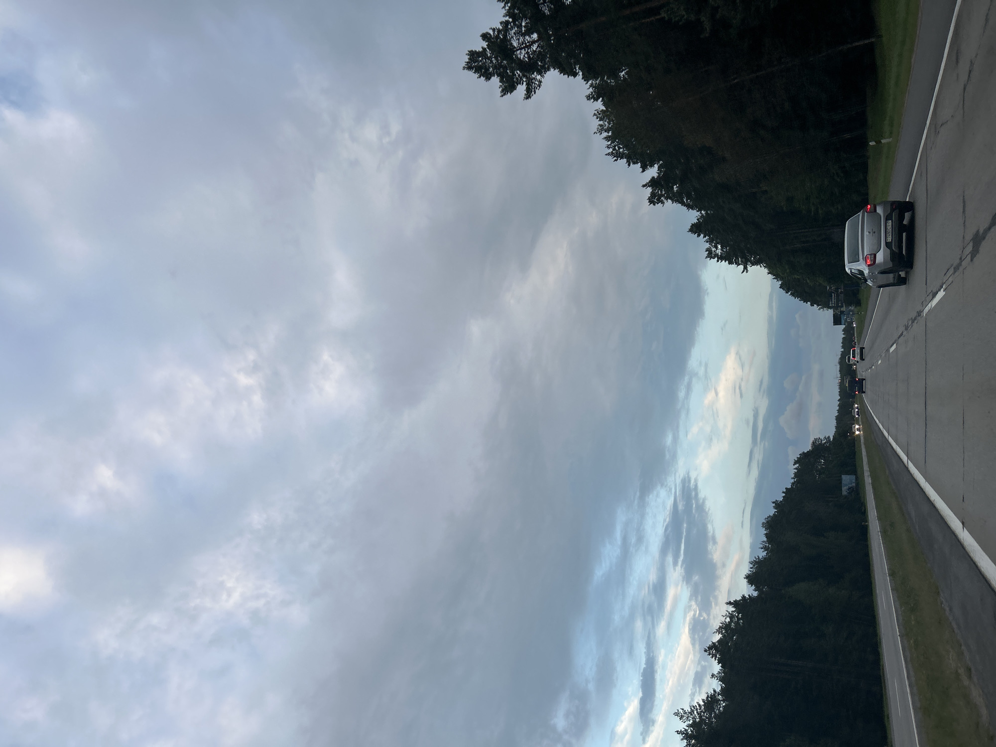 The image depicts a two-lane highway with traffic, bordered by a dense forest of pine trees on the right and a grassy shoulder on the left. The sky is overcast with a mix of gray and light blue clouds, suggesting either dawn or dusk. A silver sedan is visible in the right lane, moving away from the viewer, and other vehicles are present further down the road. The road surface shows some wear and tear, with visible lane markings and cracks. The overall scene is one of travel through a natural landscape under a moody sky.