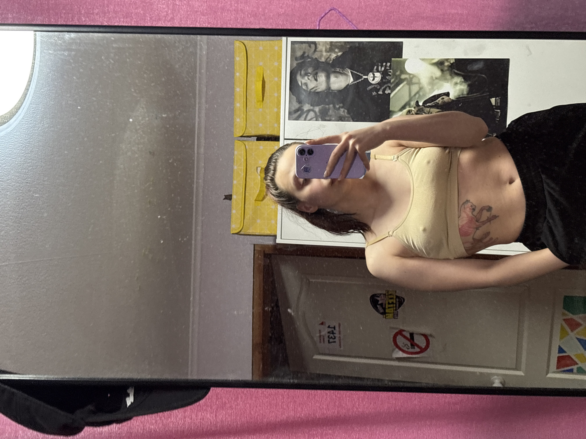 A woman is reflected in a dusty mirror, taking a photo of herself with a purple phone. She is wearing a tan-colored sports bra and black shorts. The room behind her is cluttered, featuring a yellow storage box, a framed portrait, stickers on a door, and a geometric pattern on the wall. The mirror is framed in black and resting against a pink surface. There is no information on the location of the city of Прикамский, Russia.