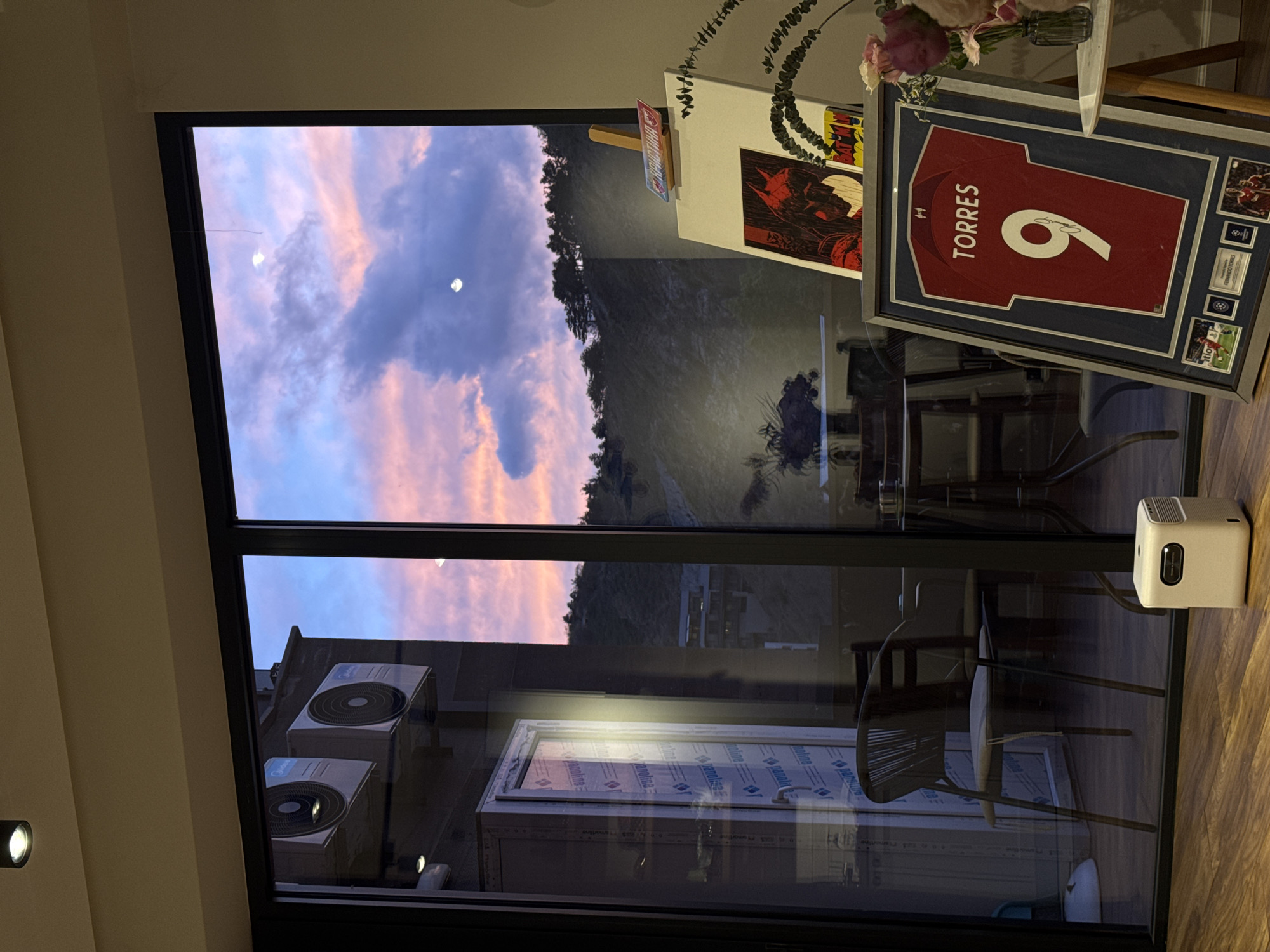 This image captures an interior view looking out a large window during what appears to be late afternoon or early evening. The sky outside is filled with soft, colorful clouds in shades of pink, orange, and purple, indicating a sunset or sunrise. A crescent moon is clearly visible against the clouded sky. Below the sky, distant hills or mountains covered in dark green foliage are visible.
Significant reflections are present on the glass window. These reflections show parts of the indoor space, including two dark chairs with woven backs, a wooden table, and a white door with "panolina" text repeated on it. Also reflected on the window, seemingly from an exterior source, are two white outdoor air conditioning units bearing the "Midea" logo, suggesting another building nearby.
Inside, to the right of the window, a framed red football (soccer) jersey is prominently displayed. The jersey has "TORRES" and the number "9" on the back, along with a team crest (likely Liverpool F.C.). The frame also contains smaller sections with text that appears to include "FERNANDO TORRES" and "CHAMPIONS LEAGUE". Above the jersey, on a shelf, there is a vase holding pink and white flowers, a decorative plant, and two framed pictures. One picture clearly features "BATMAN" text, and another small red sign on the shelf also displays "Batman". A small, white, rectangular electronic device, possibly a projector, rests on the surface to the right of the window. The floor is made of wood.
The scene conveys a peaceful and personalized atmosphere, blending a scenic outdoor view with personal interests and decor.