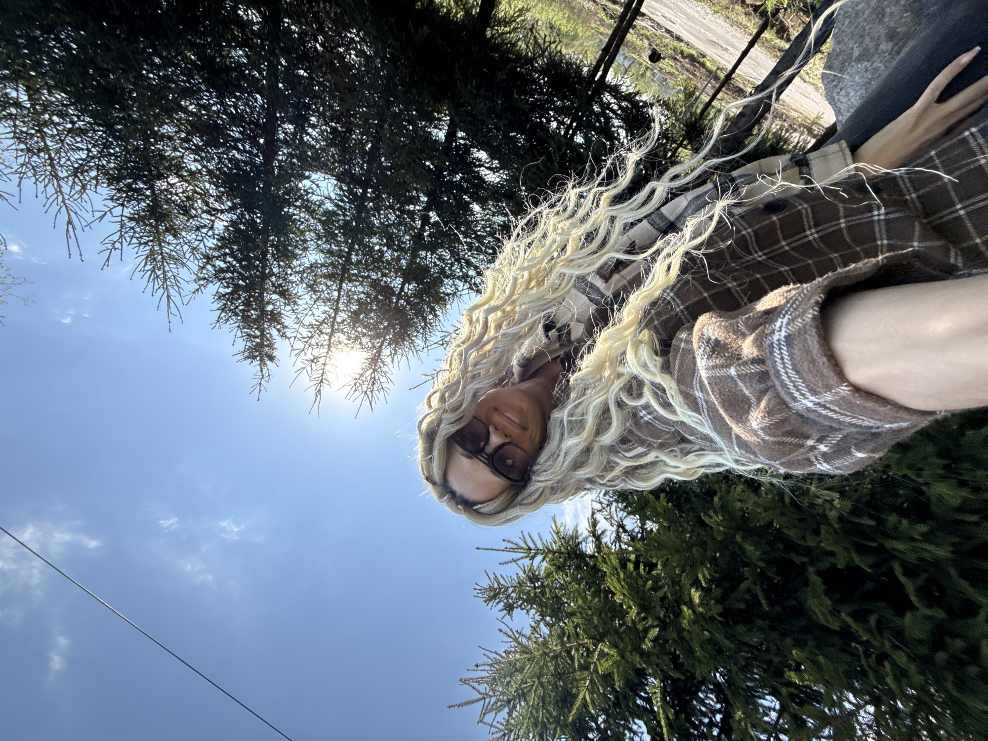 A person with long, wavy blonde hair and sunglasses is seen in a low-angle shot, wearing a brown and white plaid shirt. The background features a bright blue sky with some scattered clouds and the sun shining through the branches of evergreen trees, likely coniferous, suggestive of a wooded or park-like area. A powerline stretches across the upper left of the frame. The location could be Rusanovo, Russia, based on the request, suggesting an outdoor setting in that city.
