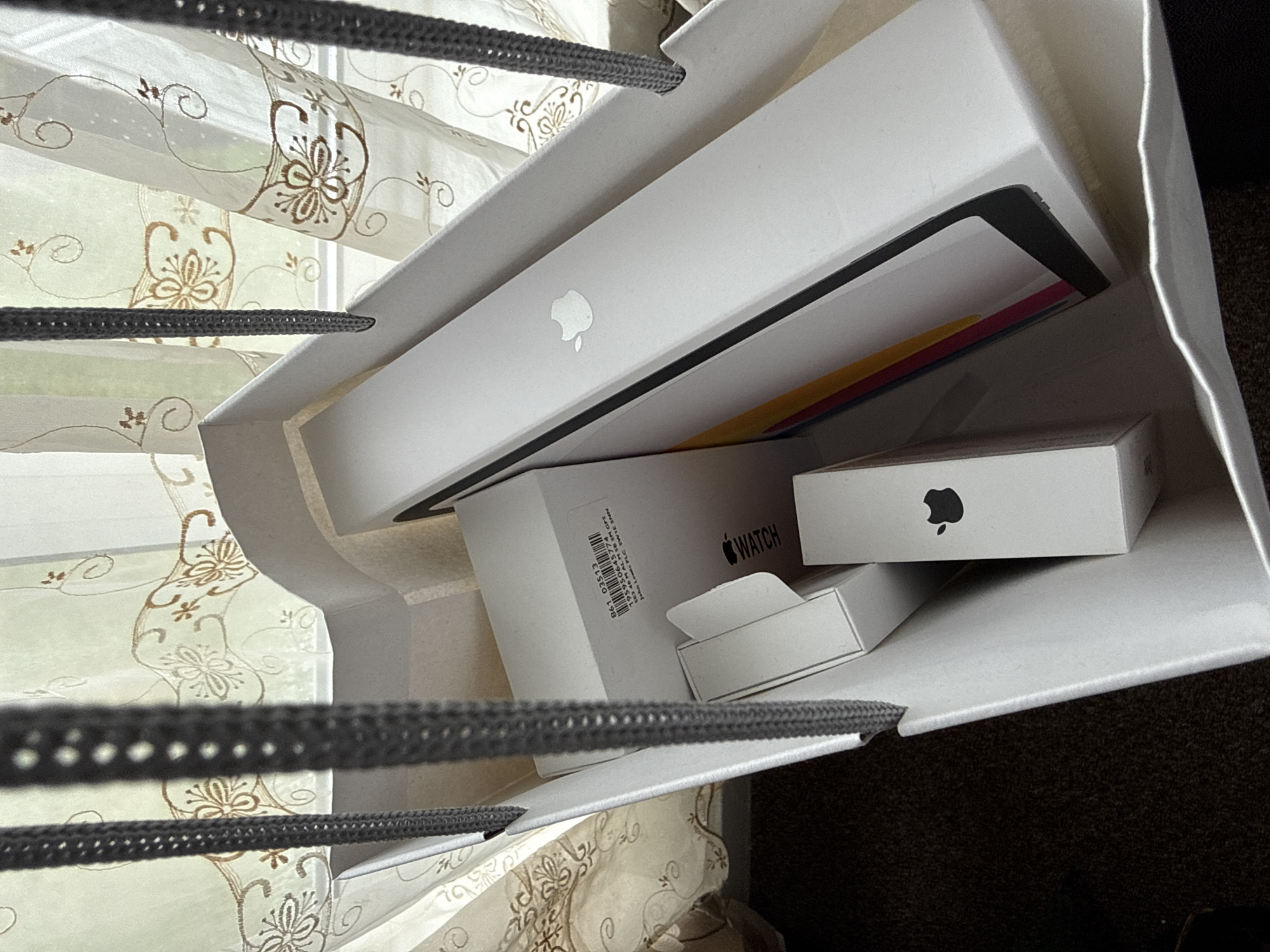 The image captures a top-down view into a white paper shopping bag filled with newly purchased Apple products. The bag has gray, braided handles visible at the top and bottom. Inside, a large white box, identified as an iPad due to its size and visible Apple logo, rests prominently. Beneath and beside it, a smaller white box labeled "WATCH" with an Apple logo is visible, indicating an Apple Watch. Another even smaller white box with an Apple logo is partially seen, likely an accessory or AirPods.
A label on the Apple Watch box displays several lines of text, including "861 03513", "195950645774", "SE3 40 MALM SB SM GPS", and "hn Lewis PLC SWIE SNN", which suggests the product's details and possibly the retailer "John Lewis PLC". Peeking from behind the iPad box are colorful edges, possibly from other product packaging.
The background, out of focus, shows light-colored curtains adorned with brown embroidered floral and swirling patterns, indicating an indoor setting, likely a room in a home, illuminated by natural daylight. The scene suggests a recent shopping trip and the arrival of new electronic devices.