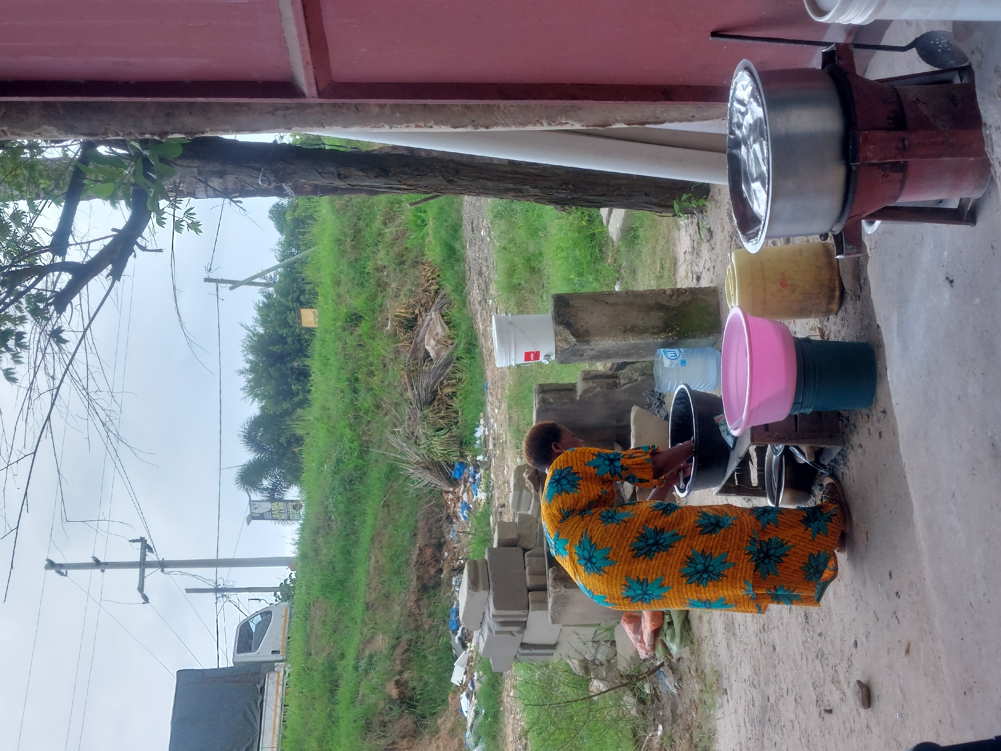 A woman wearing a yellow and turquoise floral-patterned dress is standing outdoors near several containers. She appears to be working with a large black pot next to pink and teal plastic bowls. In the background are construction materials, a grassy area, trees, and utility poles. A truck is visible in the distance. The scene appears to be in a semi-urban or rural setting. While I do not have enough information to pinpoint the exact location, the provided information mentions that the image originates from Dar es-Salaam, Tanzania.